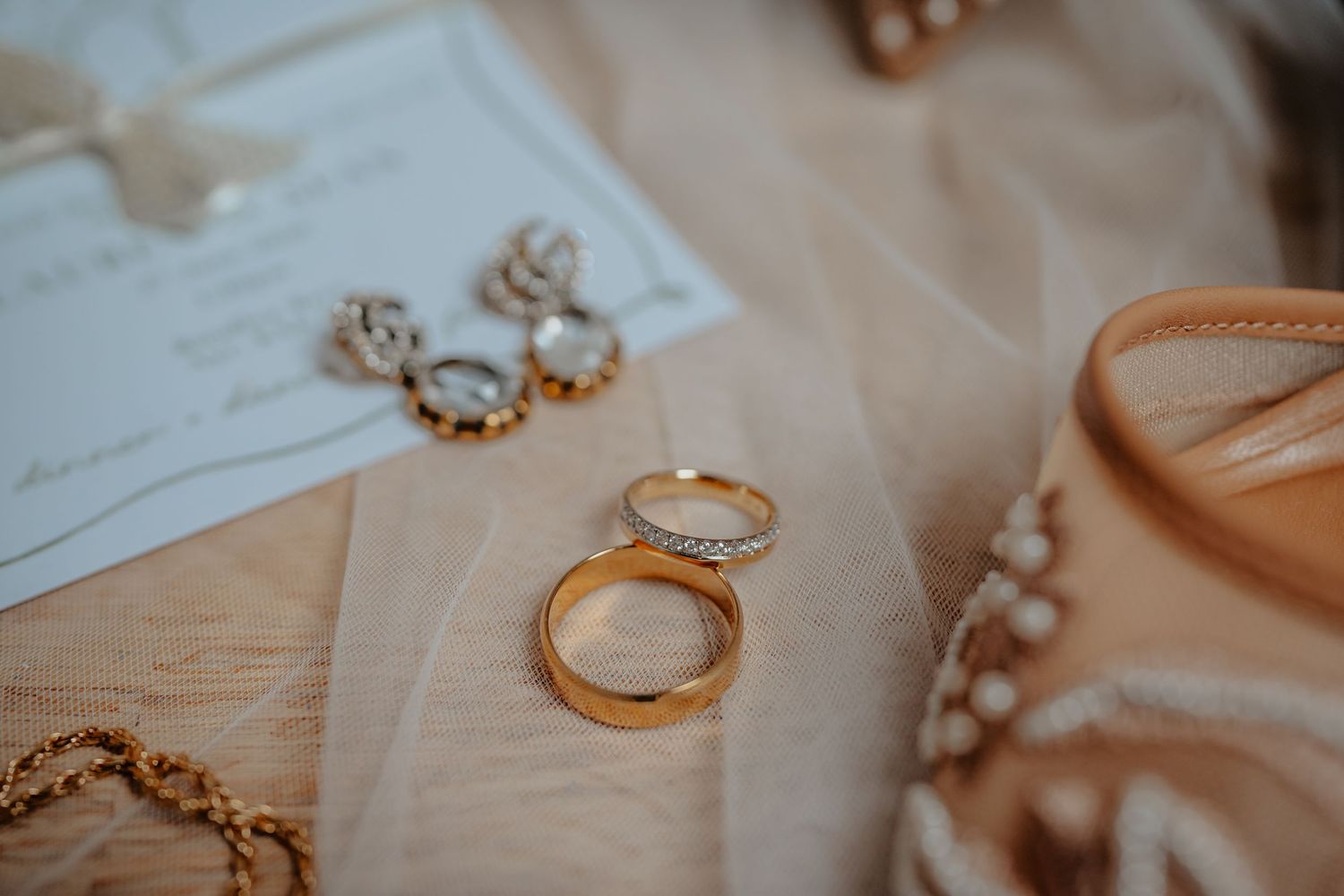 Two gold wedding bands and diamond earrings arranged elegantly on soft blush fabric beside a wedding invitation.