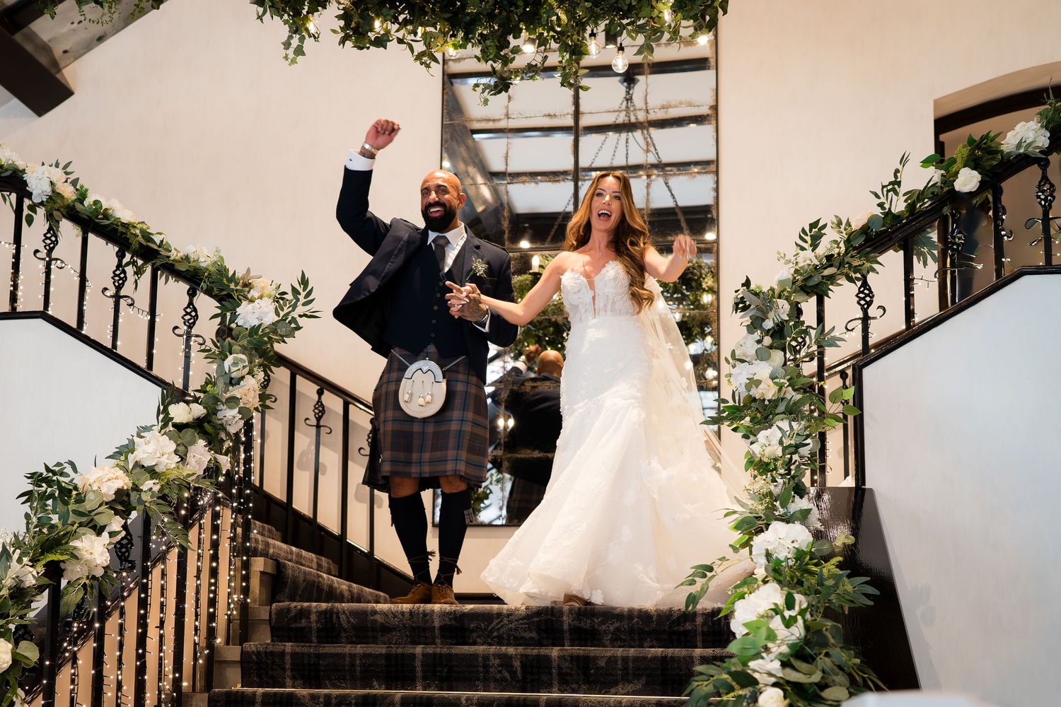 Bride in white gown and groom in Scottish kilt descend floral-adorned staircase, celebrating their wedding reception entrance.