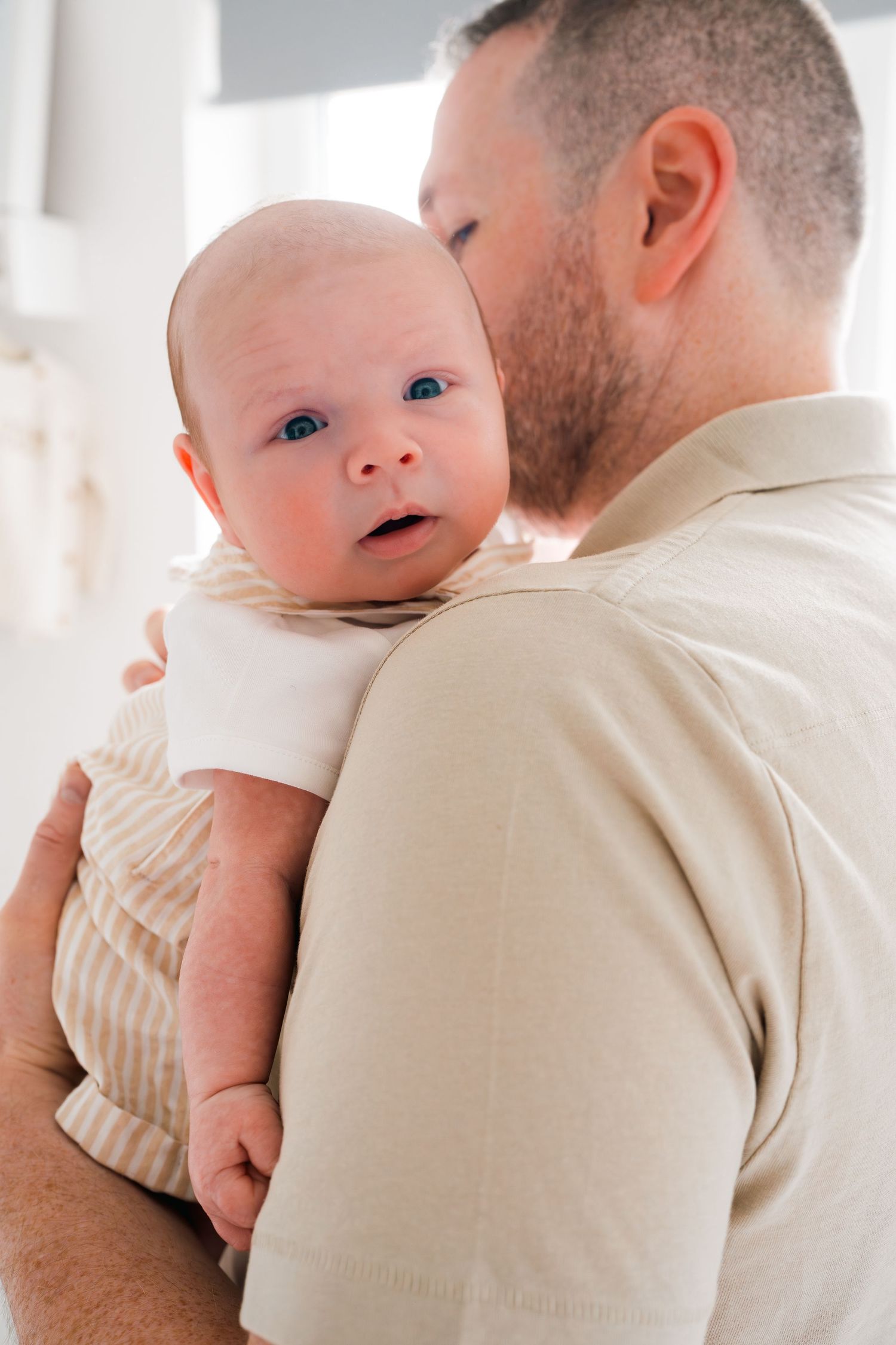 A bearded father tenderly holds his newborn baby, who gazes curiously at the camera in a bright room.