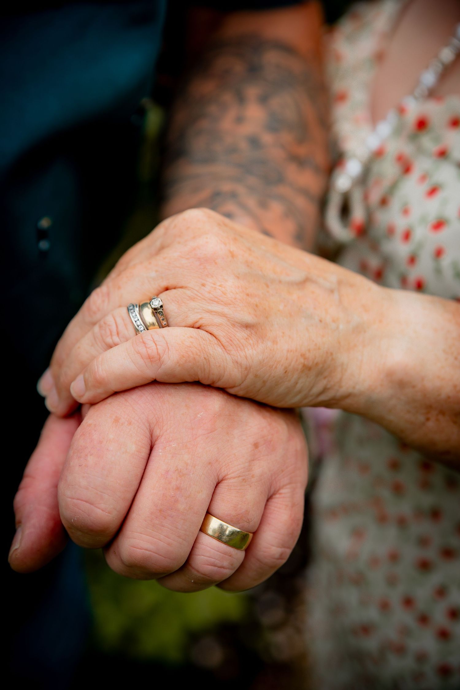 Elderly couple holding hands with wedding rings and a tattooed arm visible, symbolizing enduring love and commitment.