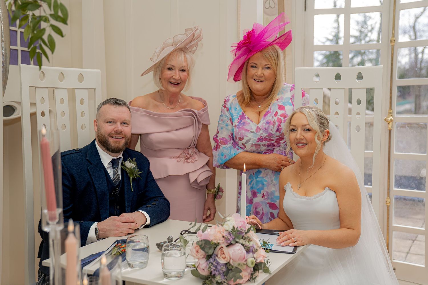 Bride and groom signing wedding register with two elegantly dressed women in pink outfits and fascinators standing behind them.