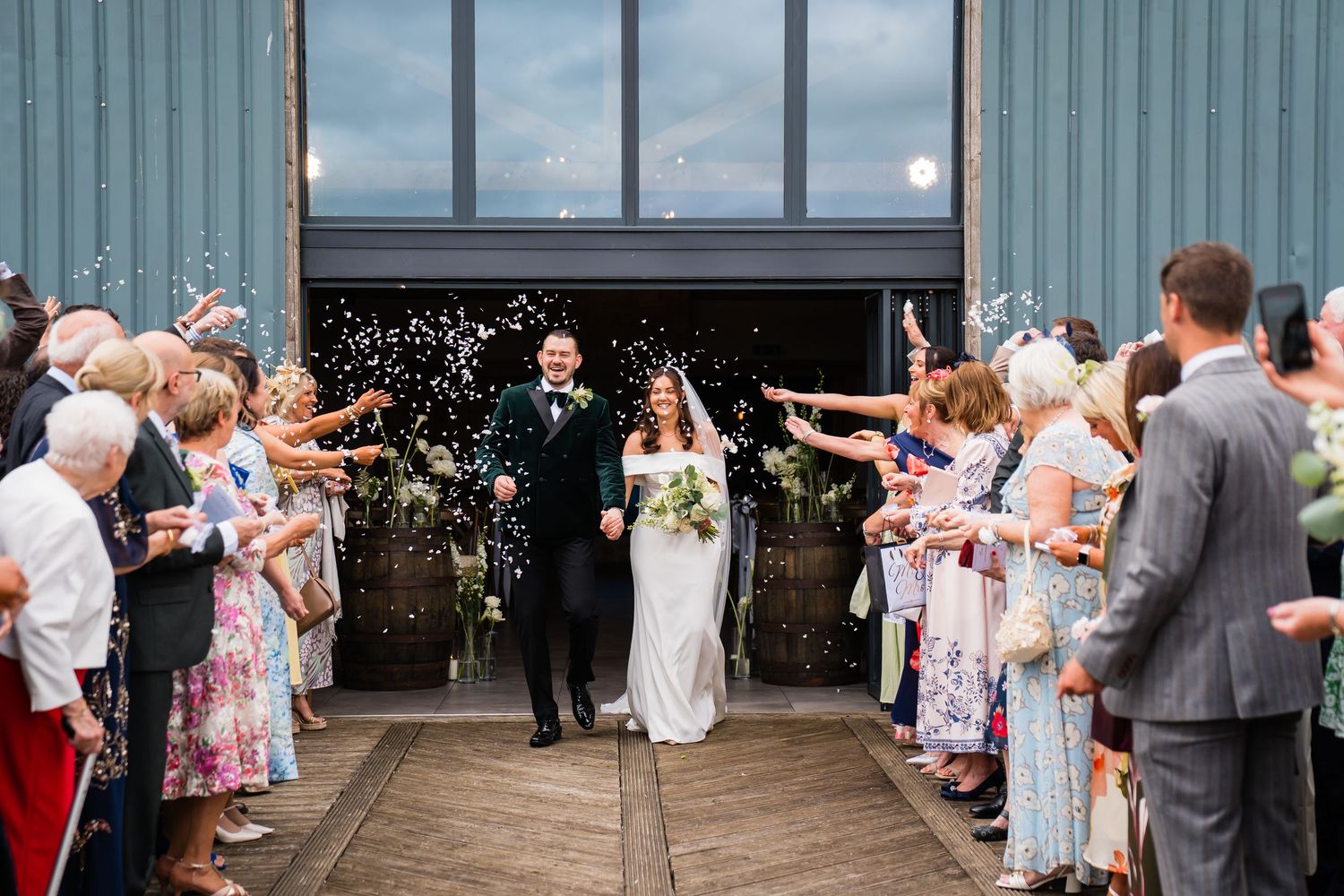 Bride and groom walk through confetti shower outside barn wedding venue, surrounded by cheering guests in colorful attire.