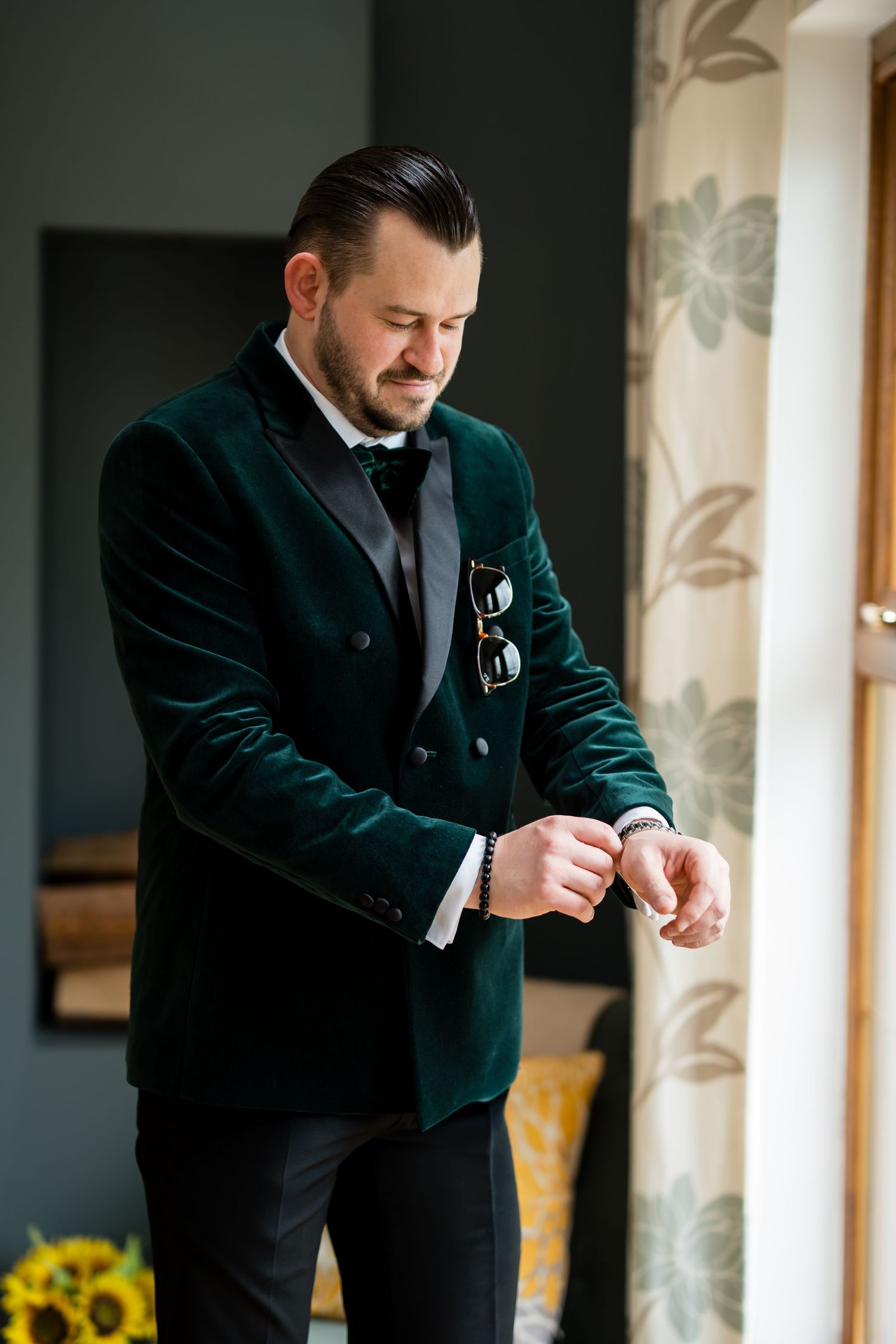 A well-dressed groom in a green velvet tuxedo jacket adjusts his cufflinks while standing near a window.
