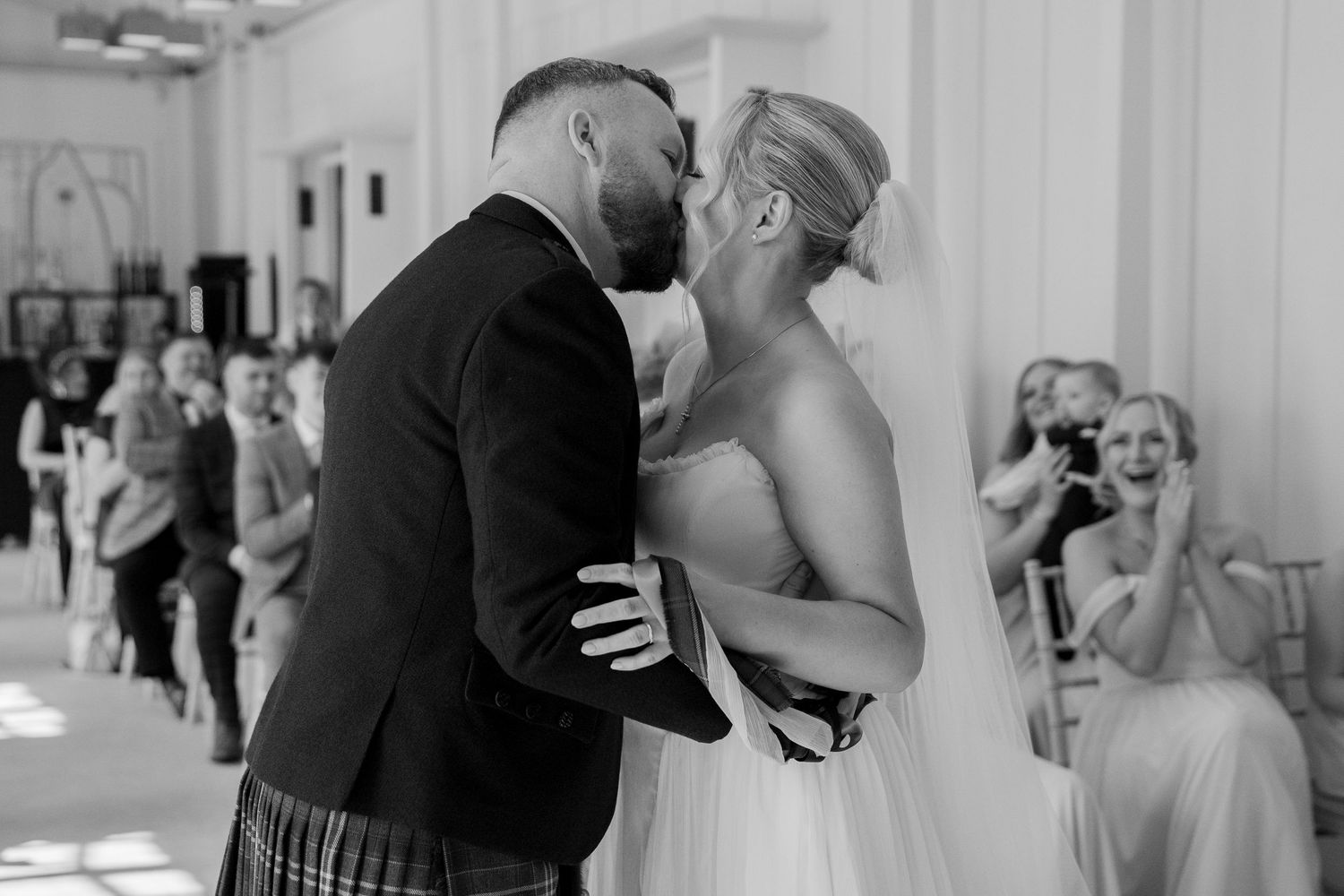 Bride and groom share a first kiss in black and white wedding photo with guests watching in background.
