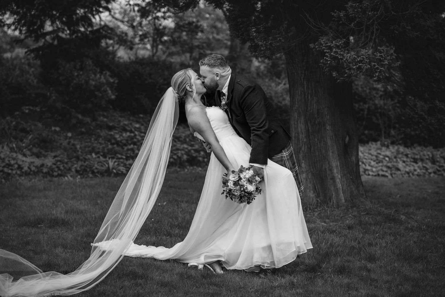 Black and white photo of a bride and groom sharing a romantic dip kiss outdoors, with a flowing veil and bouquet.