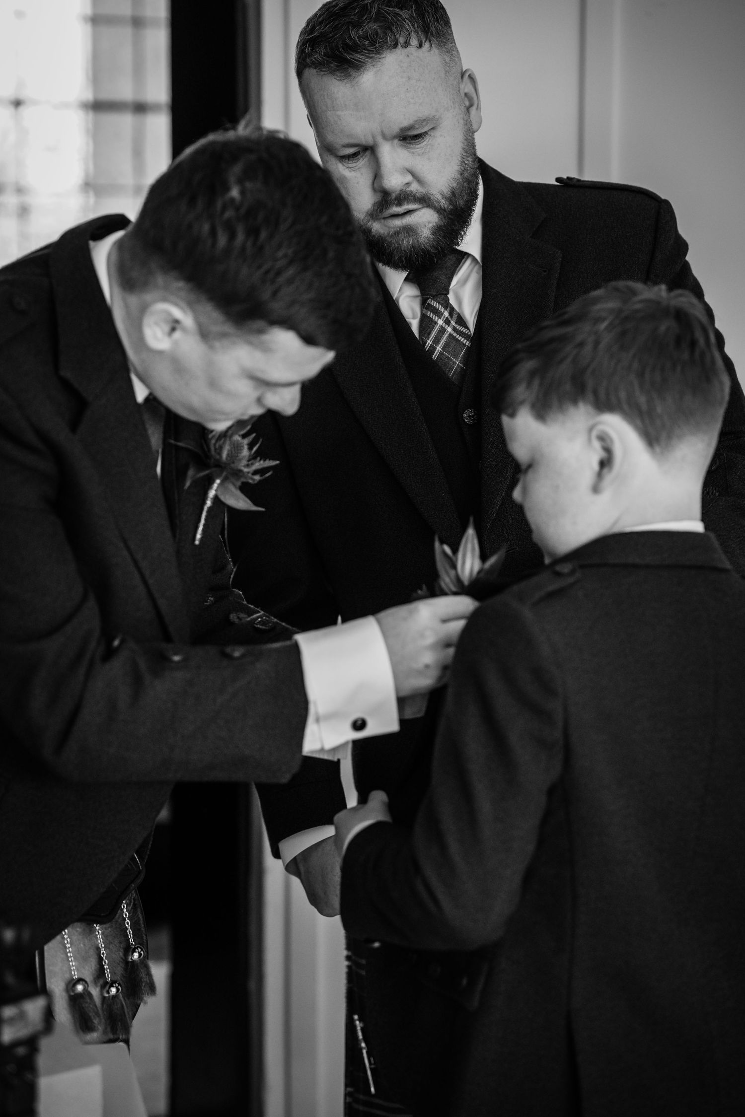 Two men in suits helping a young boy with his boutonniere at a formal wedding event, black and white photo.