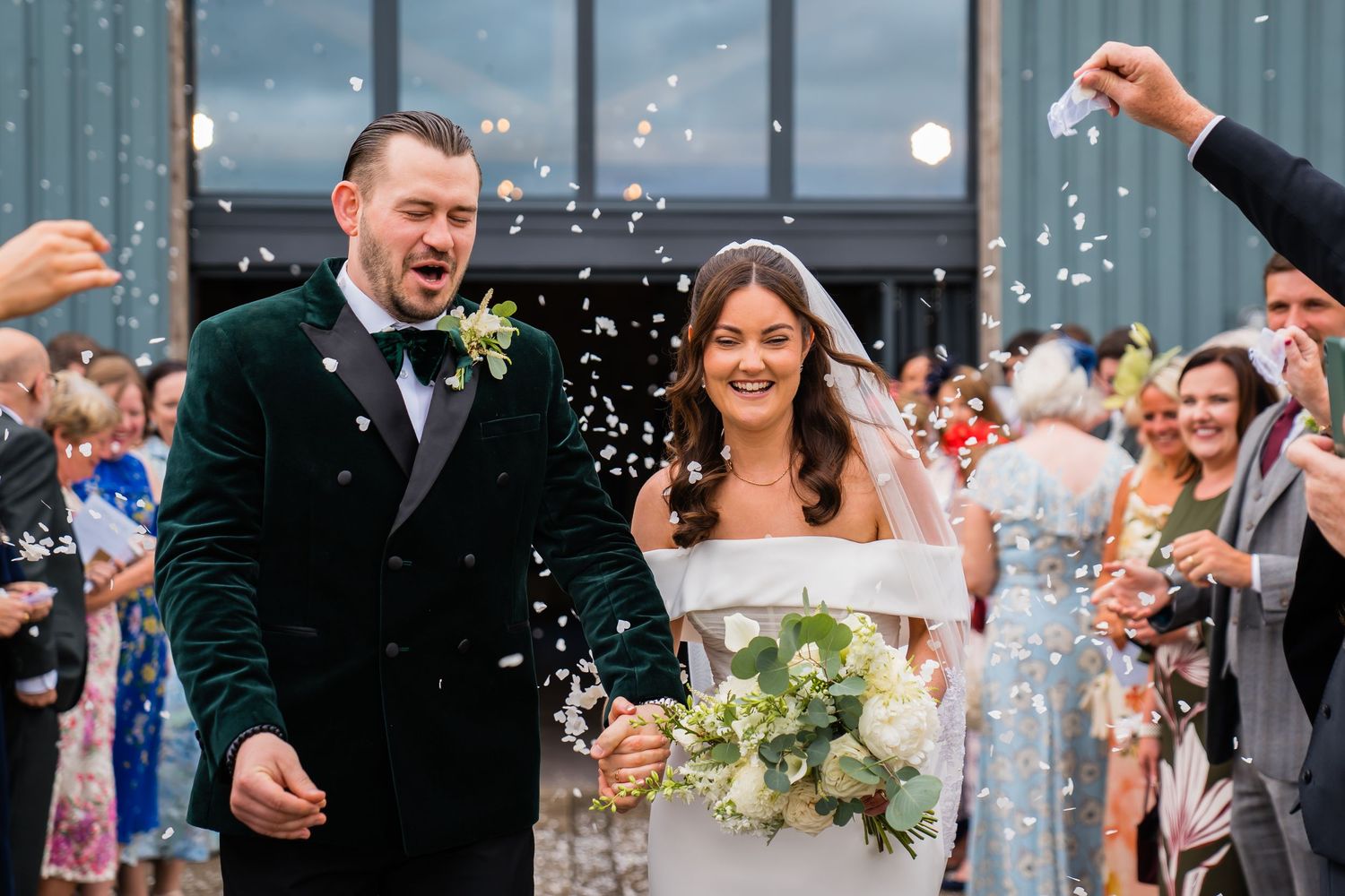 Bride and groom walk through confetti shower outside wedding venue, bride holding white bouquet, groom in green velvet suit.
