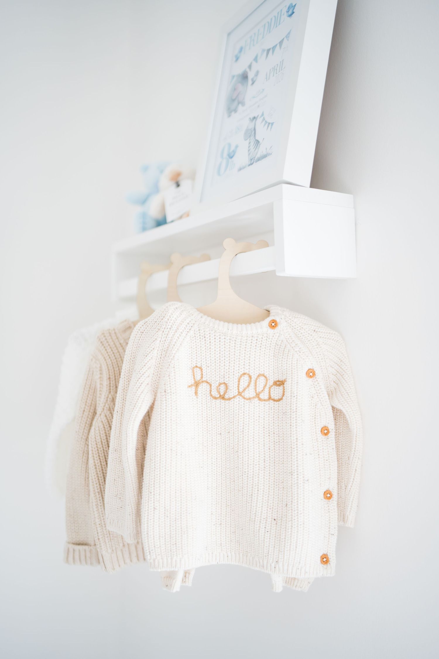 Cream knit baby cardigan with orange 'hello' embroidery hanging on a white wall shelf in a nursery.