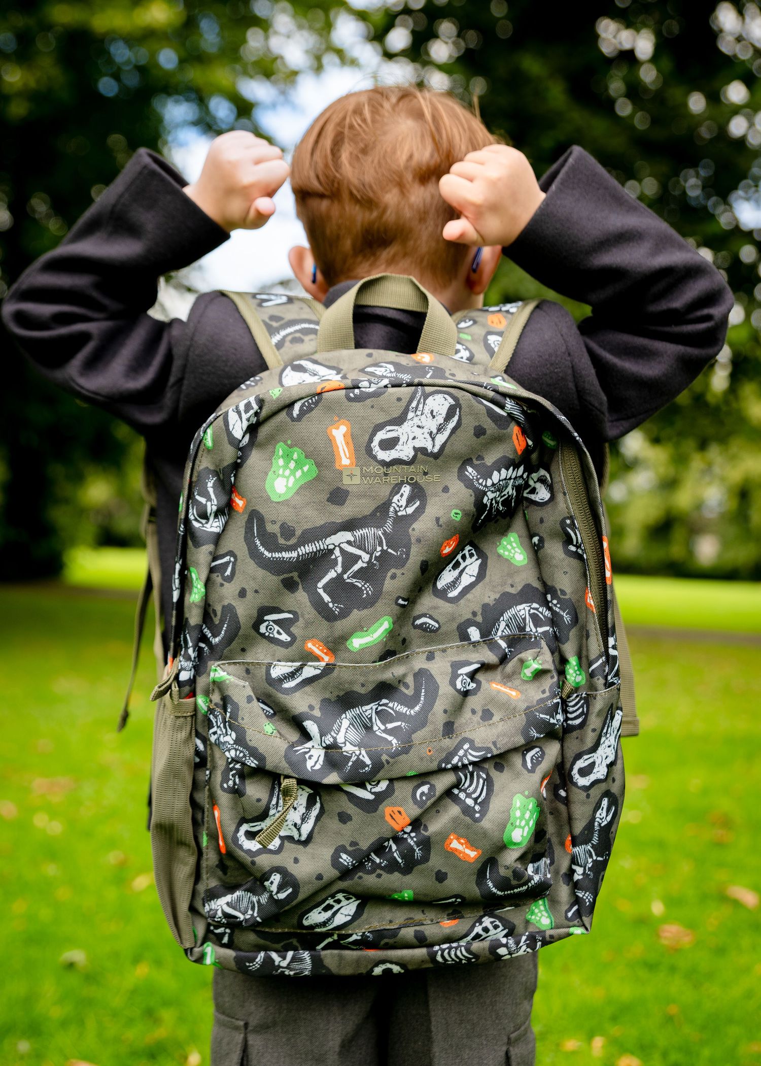 Boy wearing a dinosaur skeleton print backpack in a park, viewed from behind.