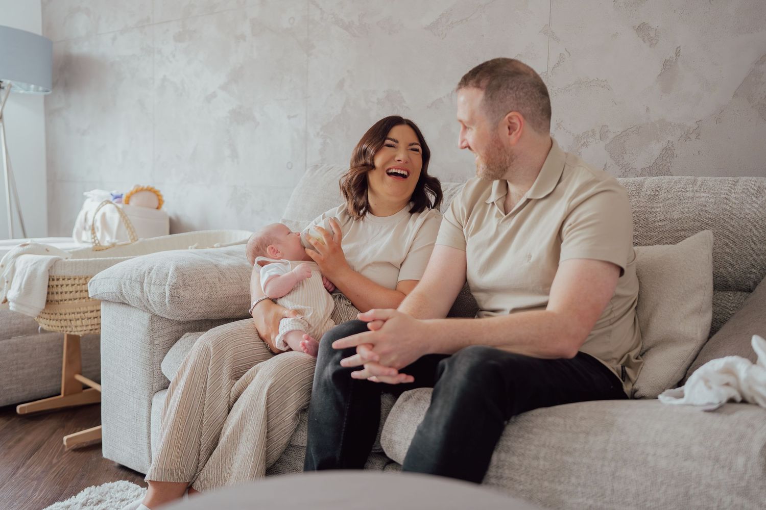 Happy couple sitting on a cozy sofa, smiling and bonding with their newborn baby in a soft, neutral-toned living room.