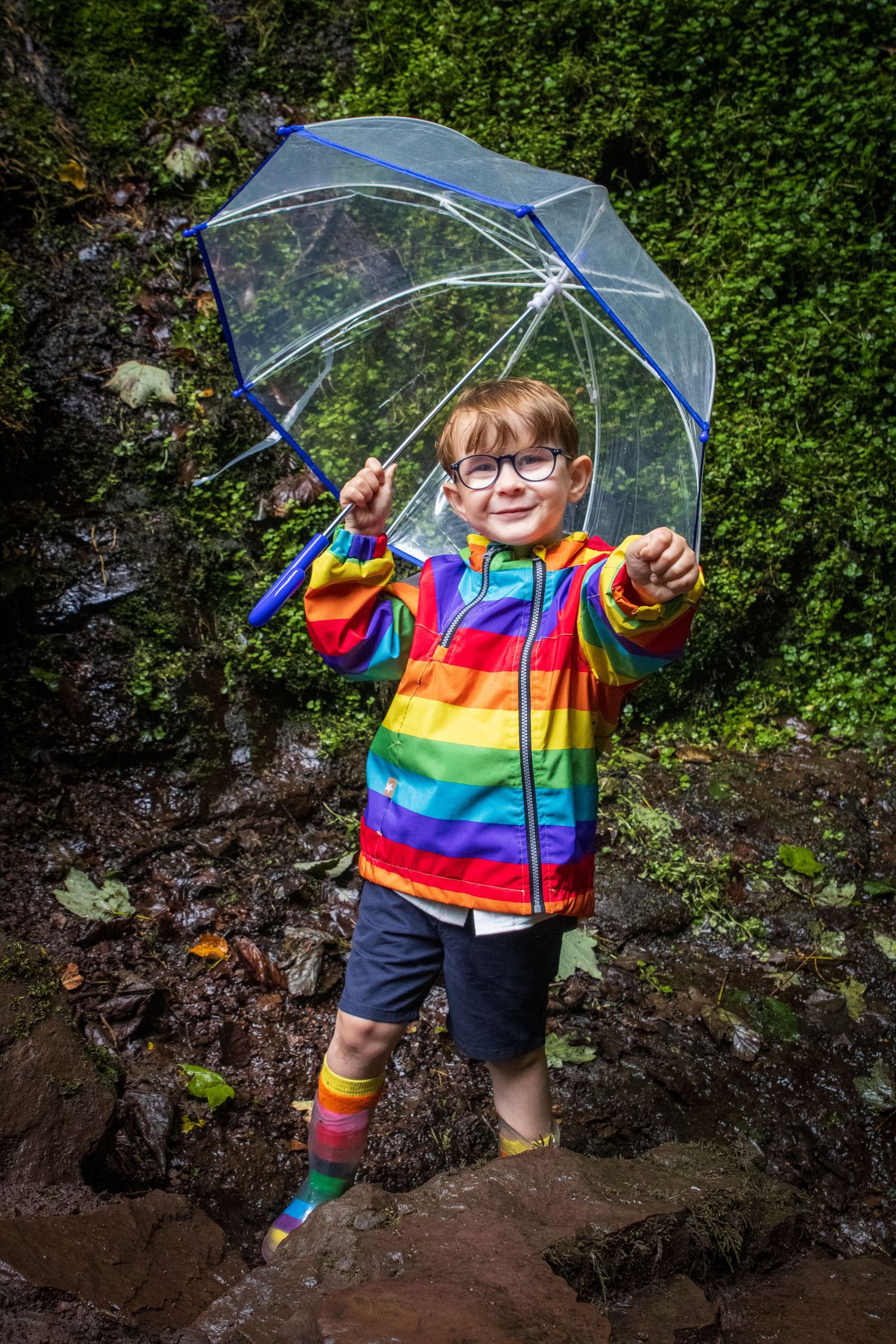 Child wearing rainbow stripes plays joyfully with clear umbrella against green foliage background.