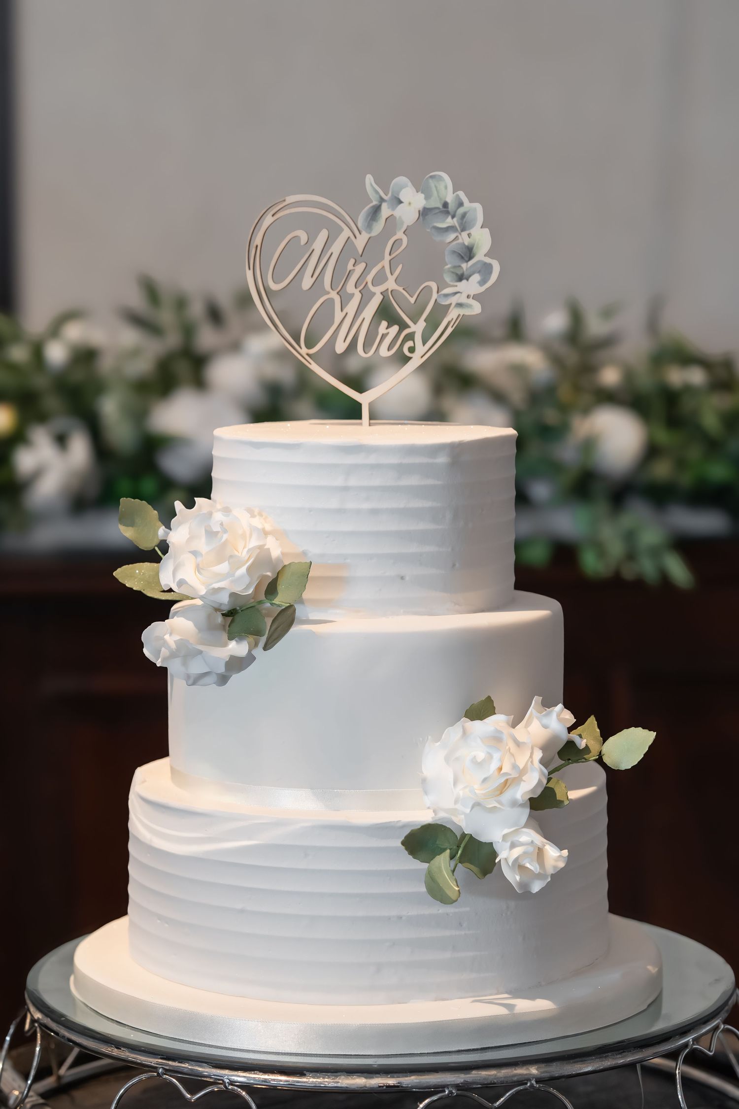 White tiered wedding cake with textured frosting, eucalyptus leaves, and white floral decorations displayed on glass stands.