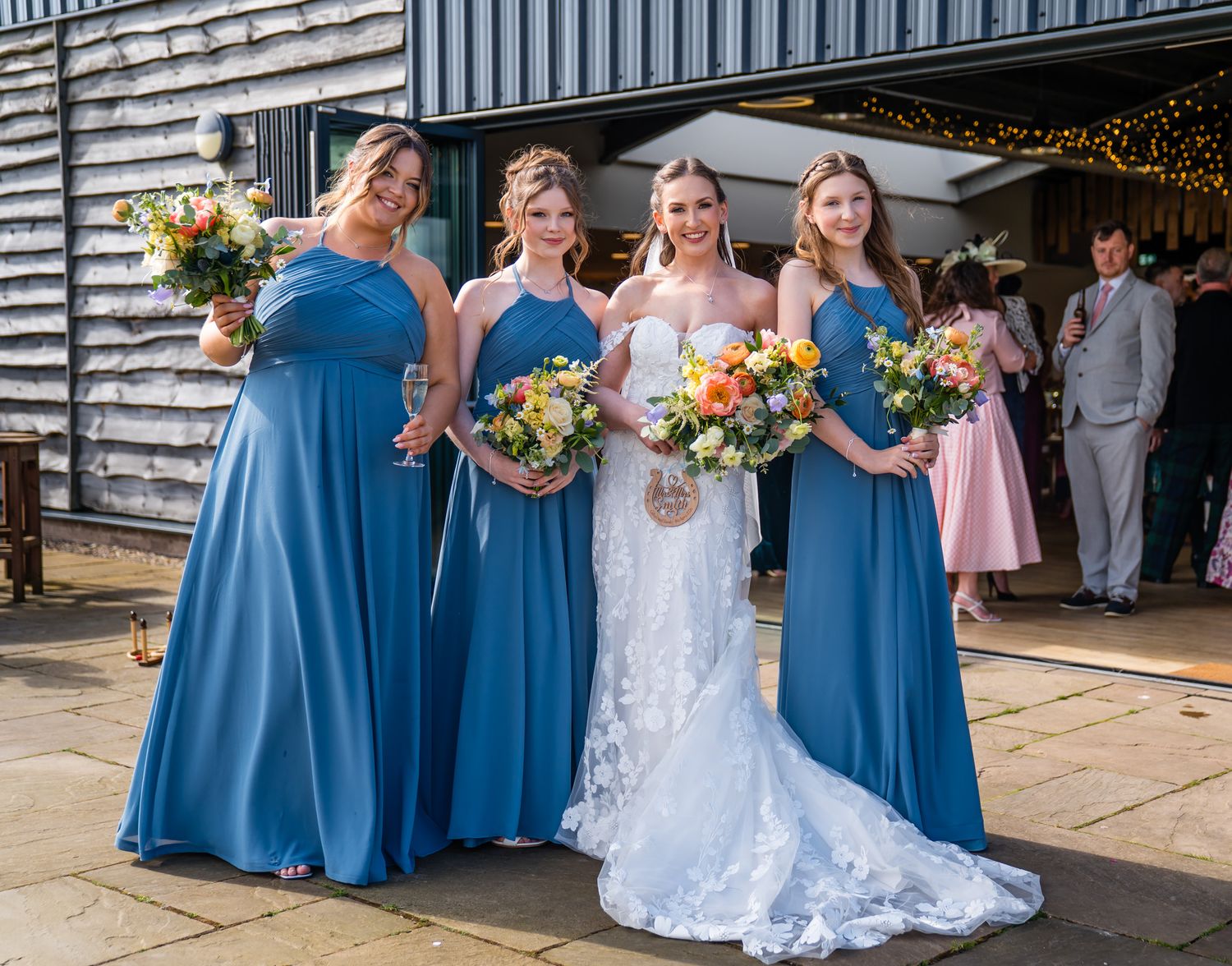 Bride in white lace gown with three bridesmaids in blue dresses holding colorful bouquets outside a rustic barn venue.