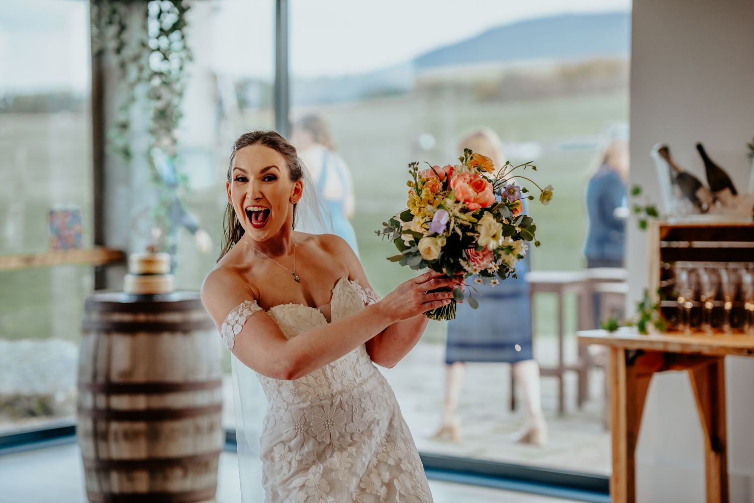 Excited bride in lace wedding dress holds colorful bouquet indoors at rustic venue with wooden barrel.