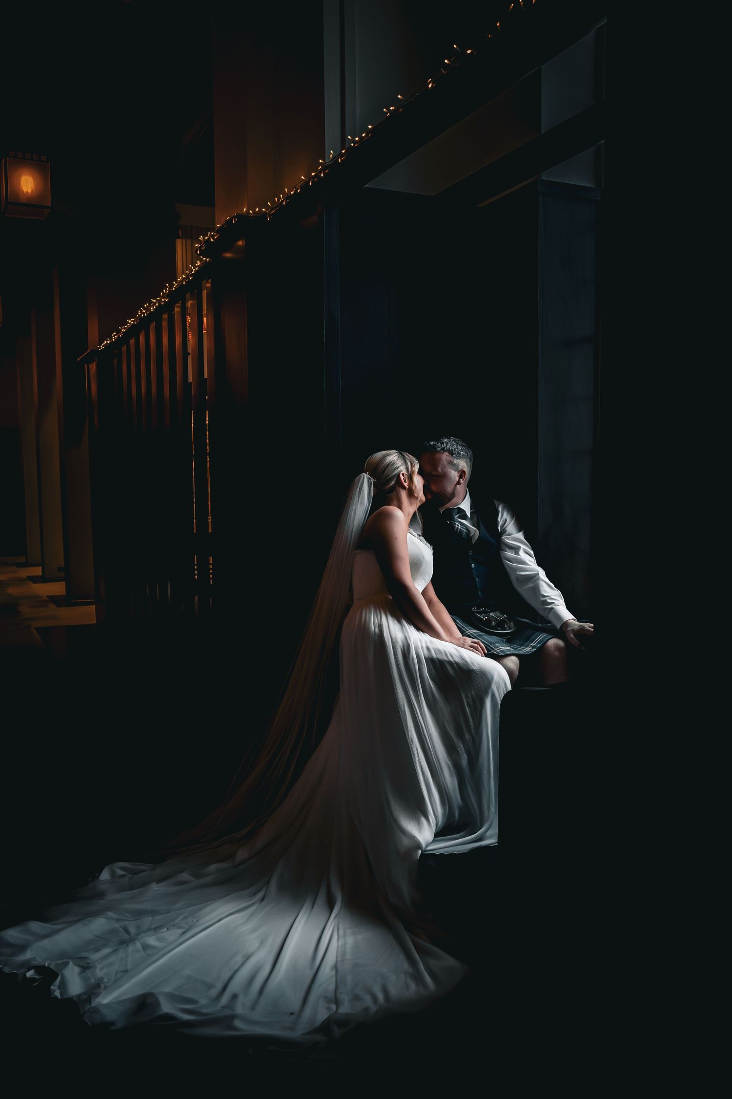 Bride in white gown and veil sits with groom in kilt on dark staircase, illuminated by fairy lights.