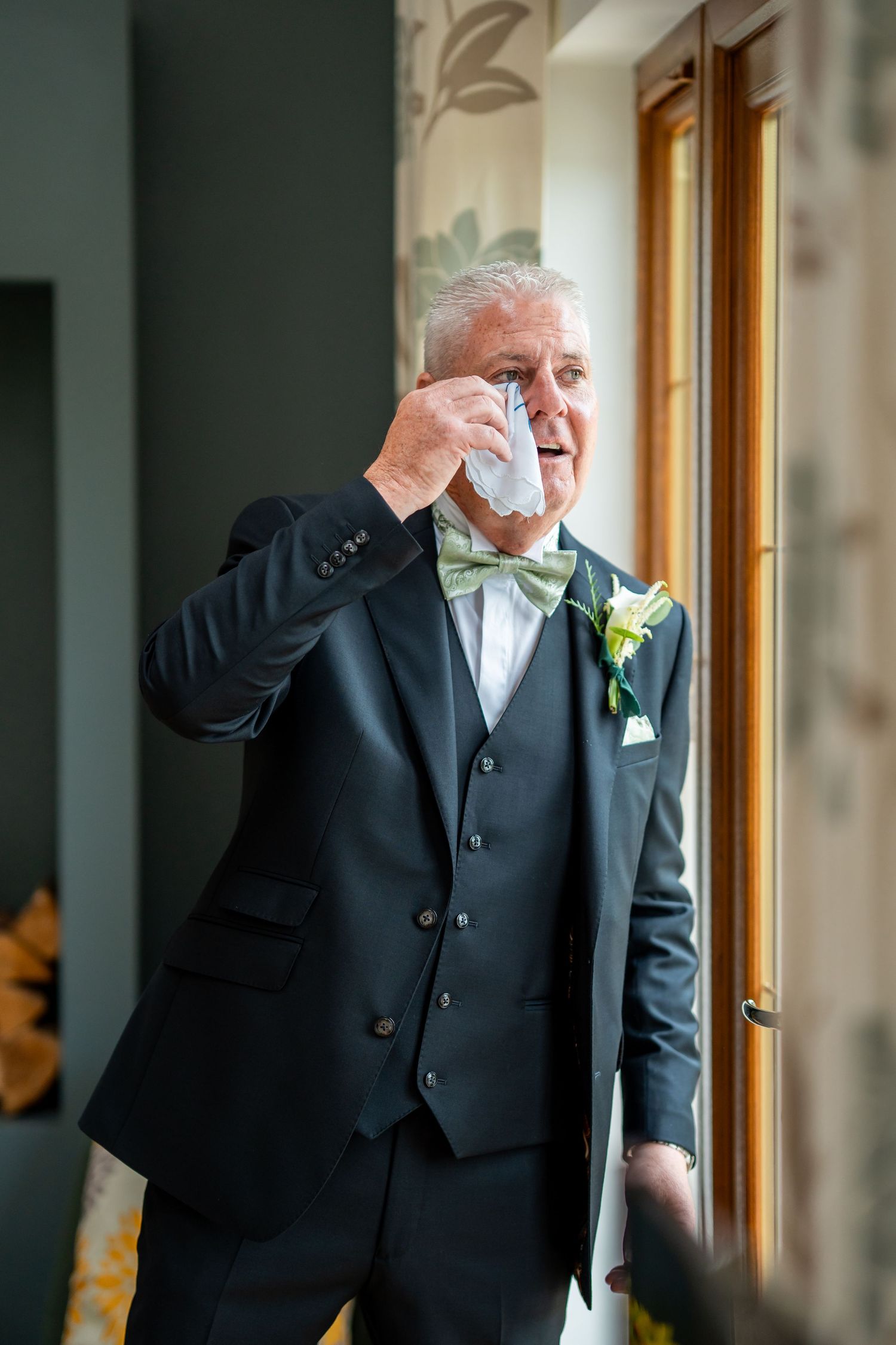 Elderly groom in dark suit with green bow tie wipes tears at wedding ceremony near window.