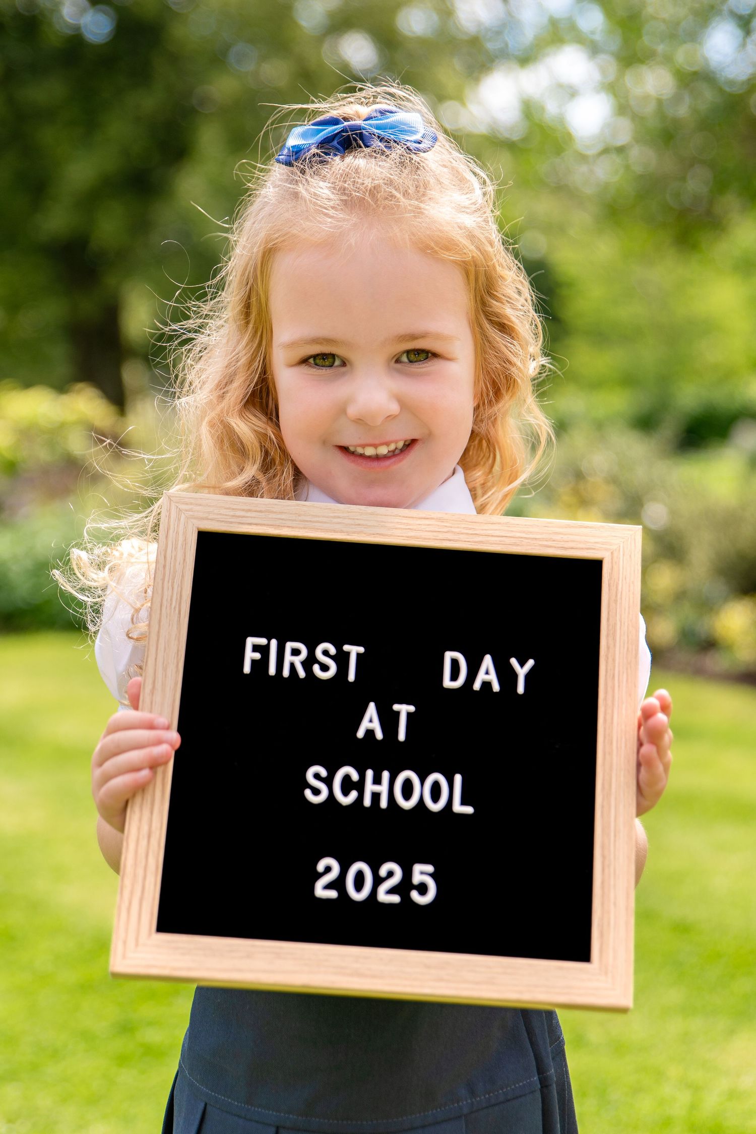 Young girl in school uniform holds a letter board sign reading First Day at School 2025 outdoors.