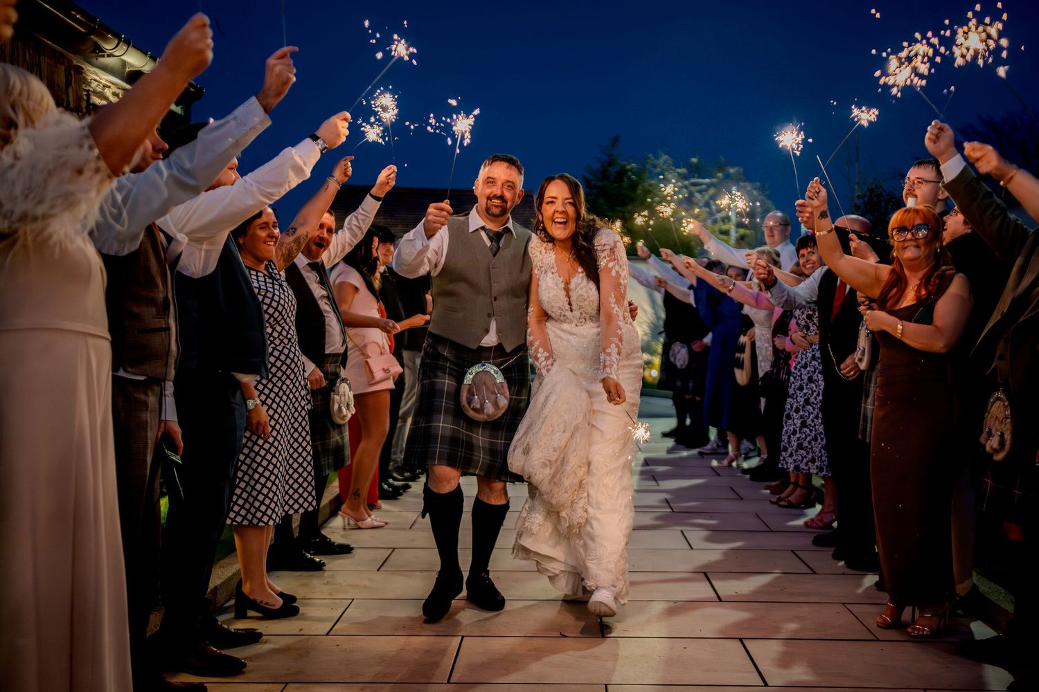 Bride and groom walk through sparkler tunnel at night wedding celebration outdoors.