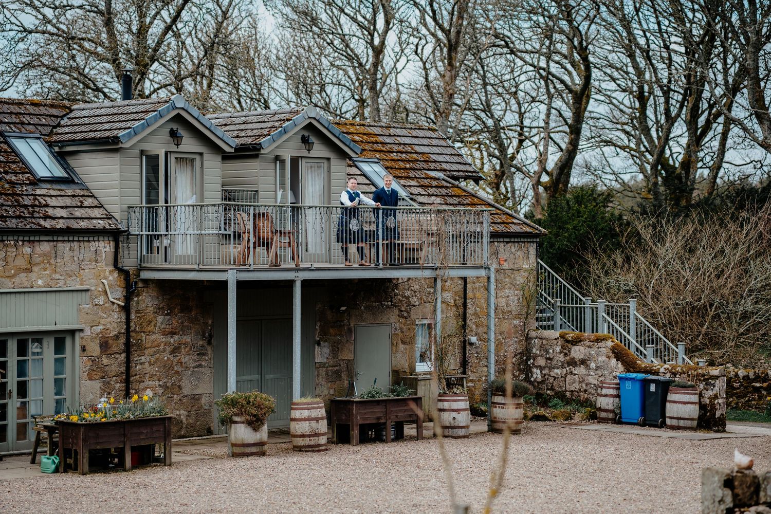 Rustic stone cottage with balcony, wooden barrels, and bare winter trees in a charming countryside setting.