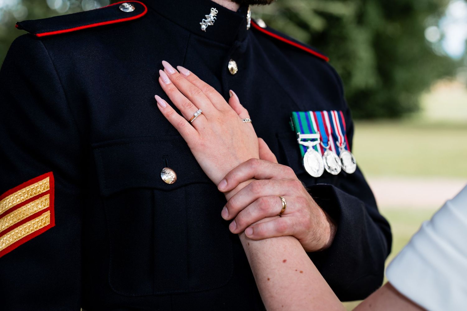 Military officer in dress uniform with medals holds hands with partner wearing engagement ring outdoors.