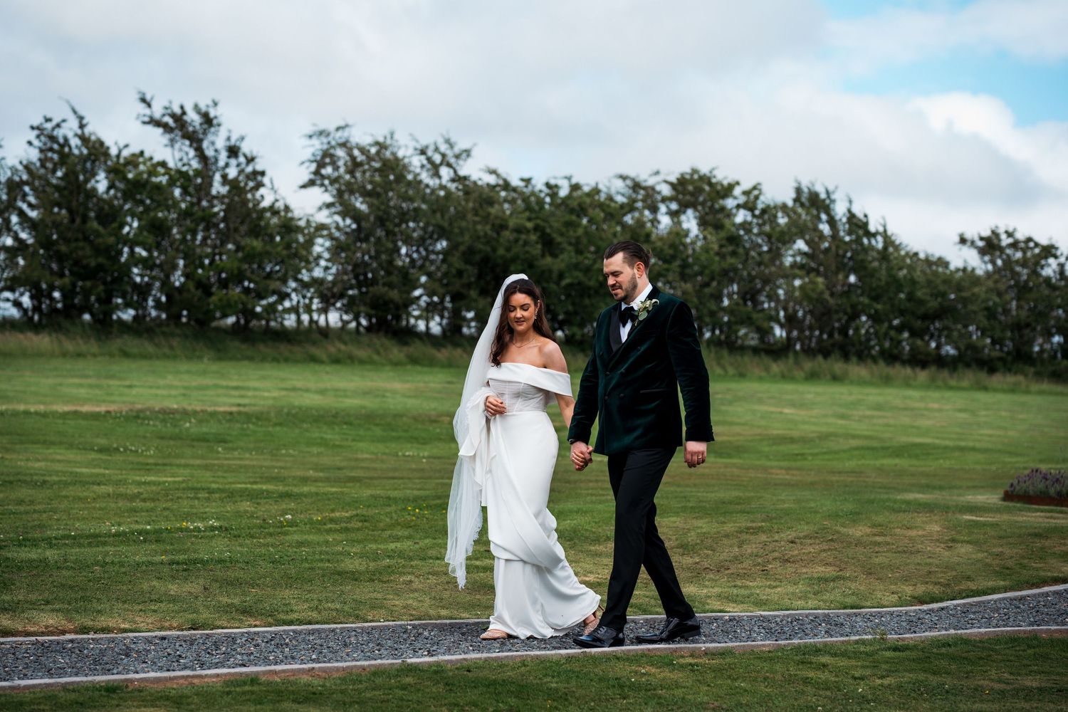 Bride and groom walk hand in hand across lush green lawn in elegant wedding attire on a cloudy day.
