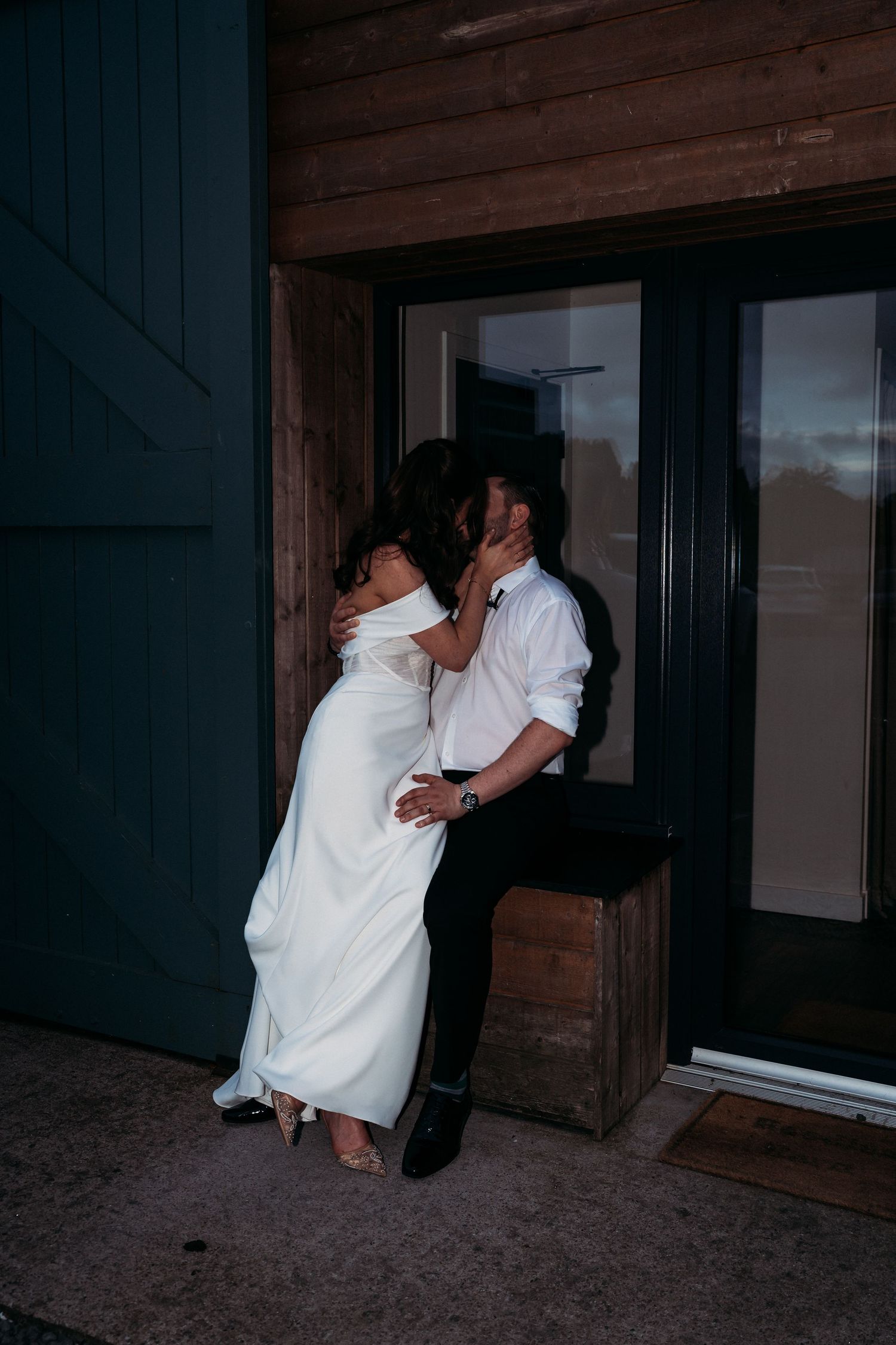 A bride and groom share a romantic kiss outside a rustic wooden venue at night, dressed in elegant white wedding attire.