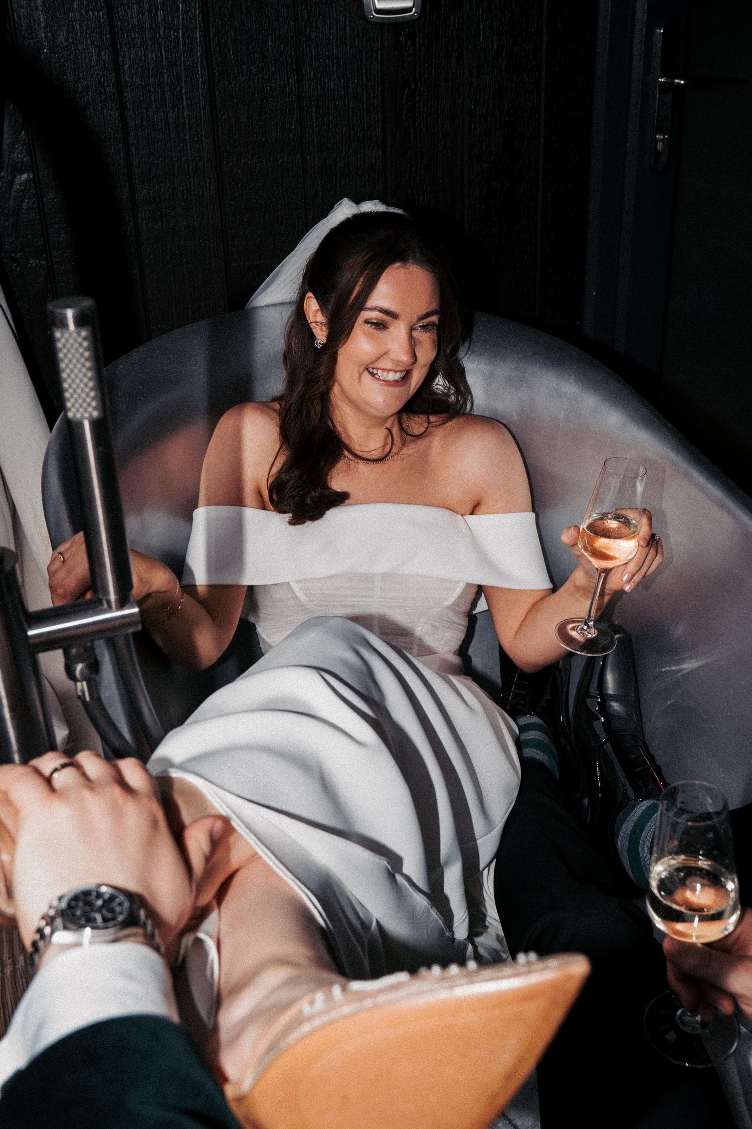 A smiling bride in a white off-shoulder gown relaxes in a tub chair, holding a glass of rose wine at her wedding reception.