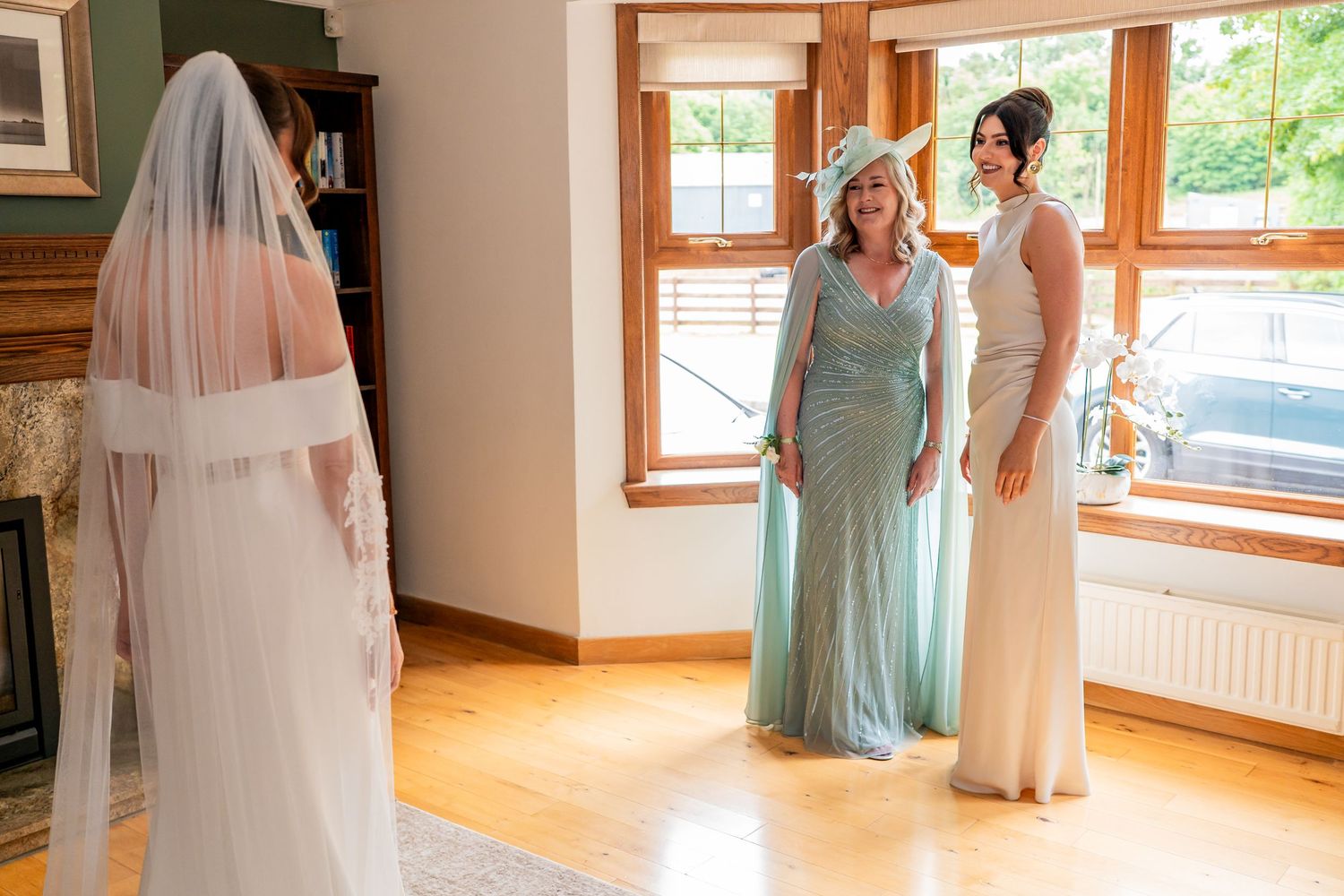 Bride in white gown and veil stands before two women in elegant dresses during wedding preparations indoors.