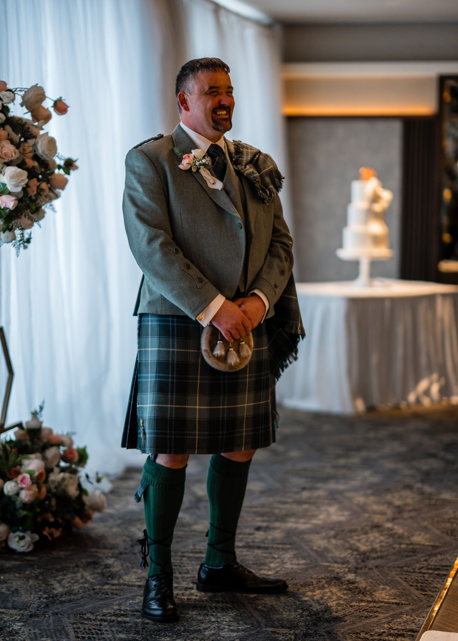Man in traditional Scottish Highland dress with green tartan kilt and jacket standing at a wedding reception.