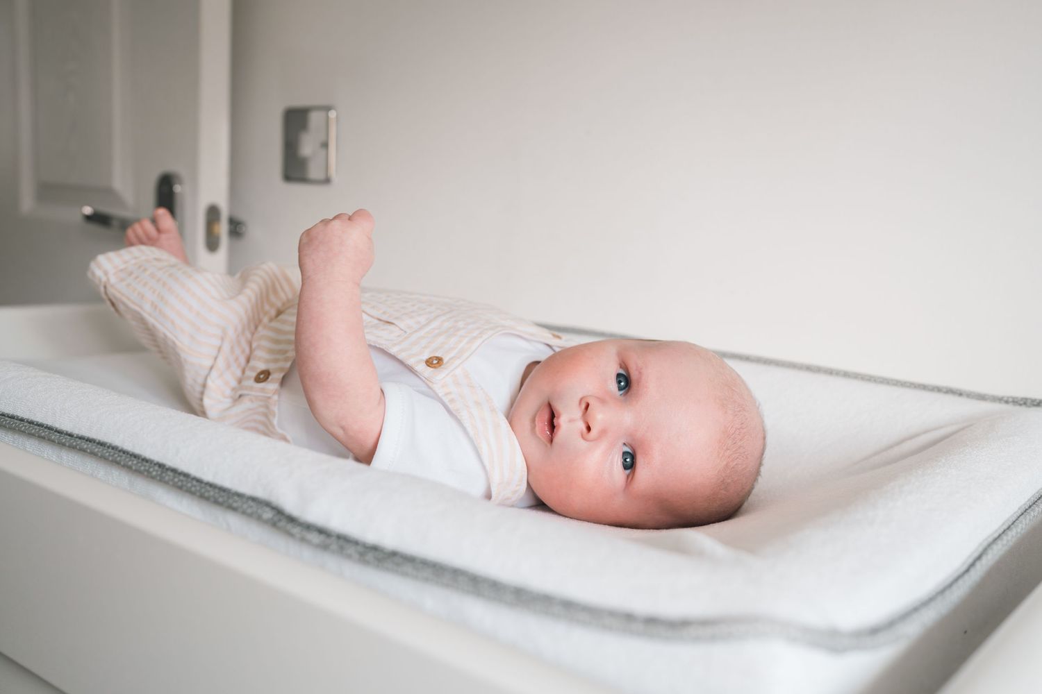 Adorable baby lying on a white changing table wearing a pink knit outfit, looking up with bright curious eyes.
