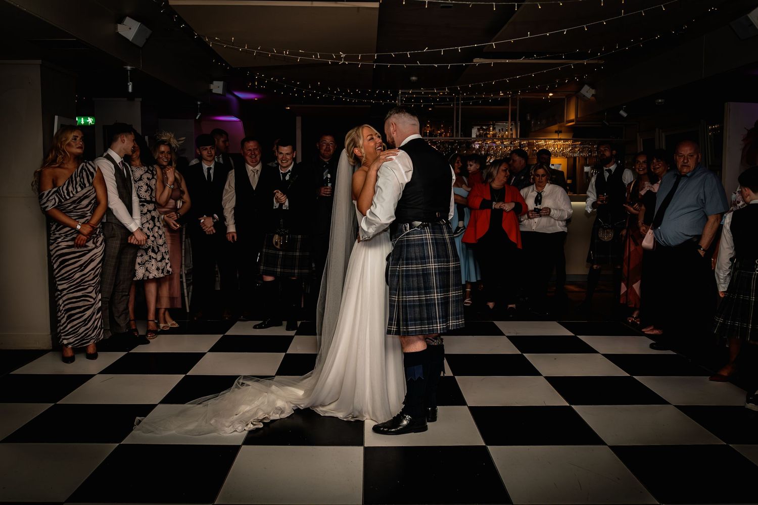 Bride and groom share first dance on checkered floor, guests watching, groom in Scottish kilt, bride in white gown.