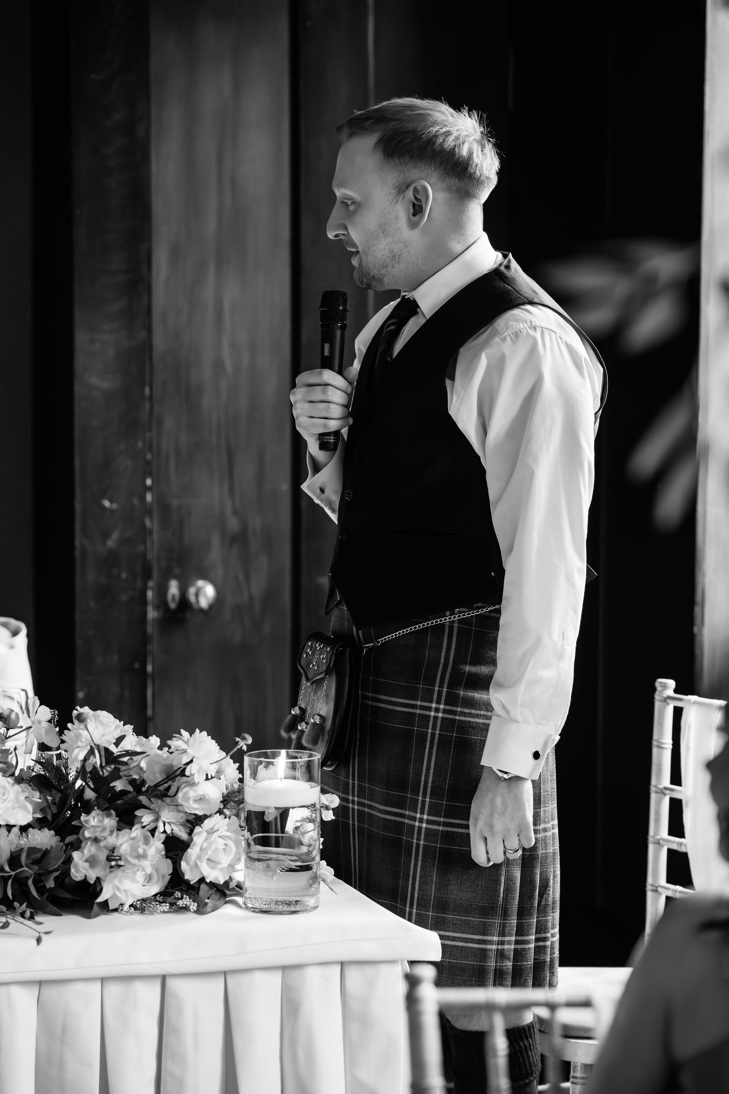 Black and white photo of a man in a kilt and vest giving a speech at a wedding reception.
