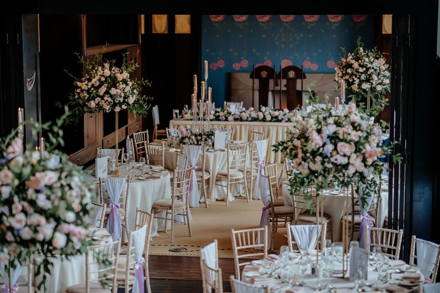 Elegant wedding reception hall with floral centerpieces, chiavari chairs, and pink roses on white linen tables.