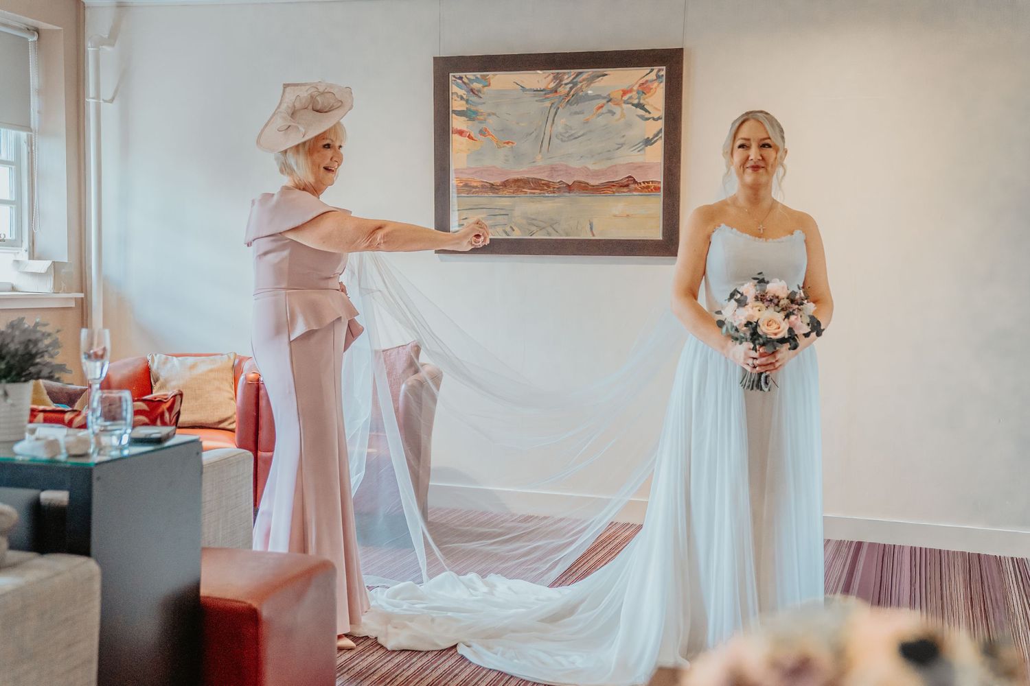 Bride in strapless gown holds bouquet as woman in pink adjusts long wedding veil in bright hotel room.