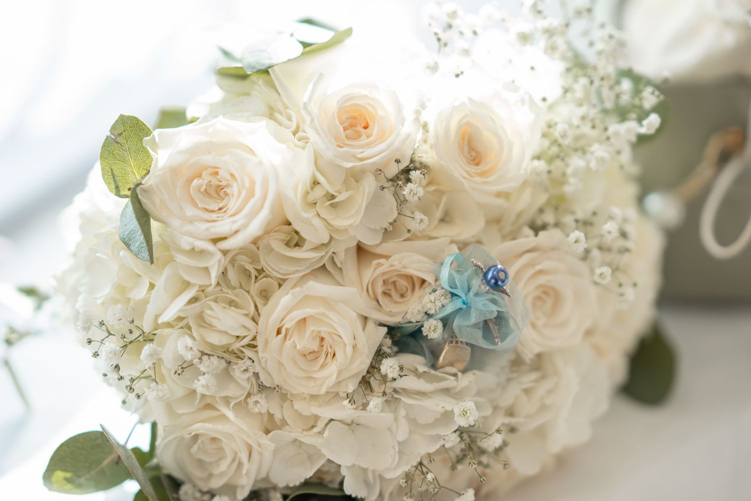 Elegant bridal bouquet featuring cream roses, white hydrangeas, baby's breath, and blue accents with green eucalyptus leaves.