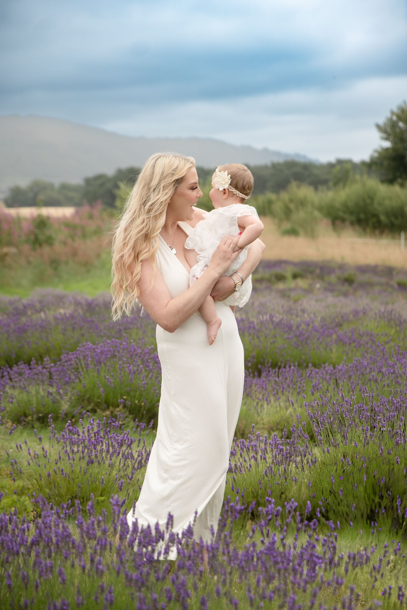 A blonde woman in a white dress holds her baby in a blooming lavender field with mountains in the background.
