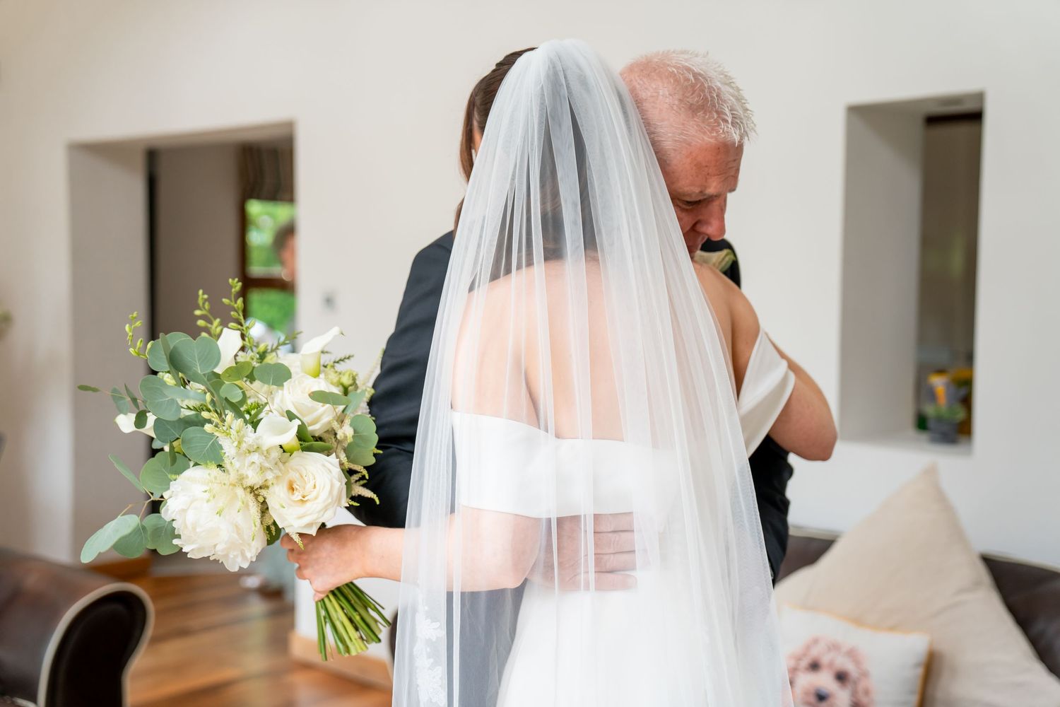 Bride in white gown and veil holding floral bouquet embraces older man in black suit indoors.