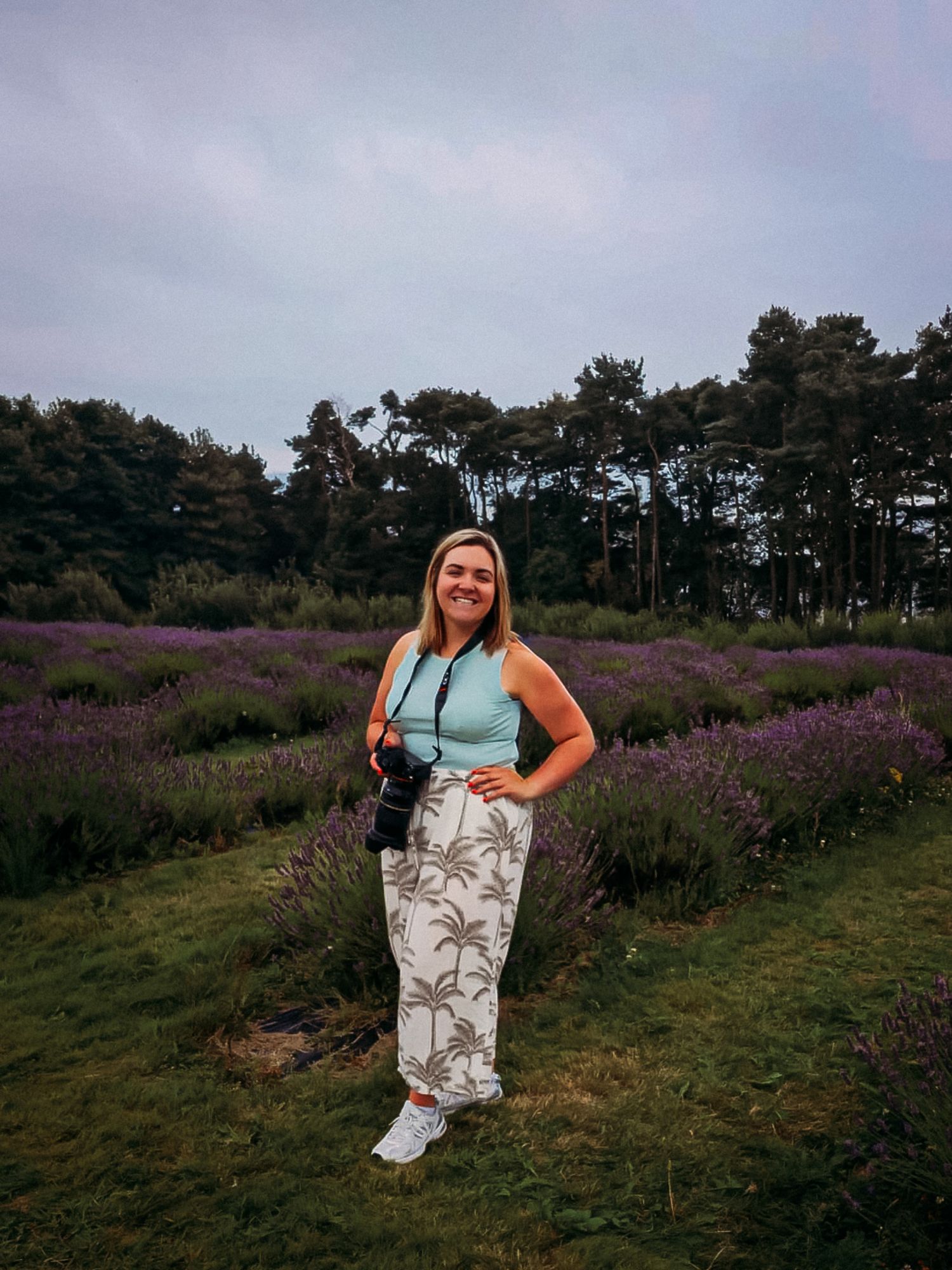 Woman in light blue top and palm print pants stands smiling in a lavender field with trees in the background.