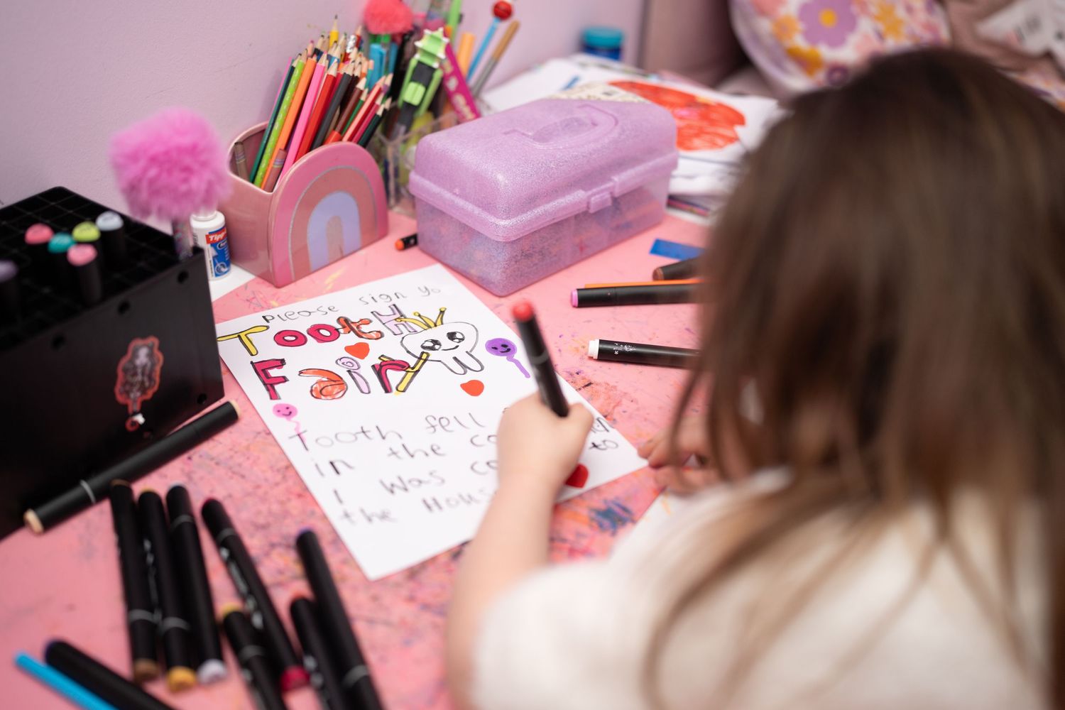 Young girl coloring a Tooth Fairy alphabet chart at a pink desk surrounded by markers and art supplies.