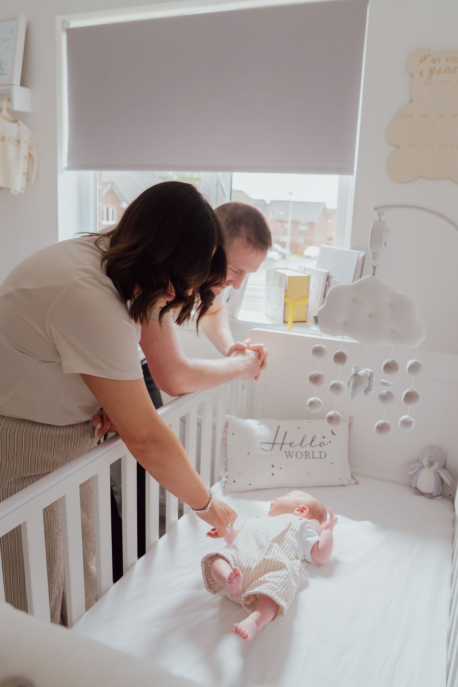 Mother leaning over white crib tending to newborn baby in knit outfit, Hello World nursery pillow visible.