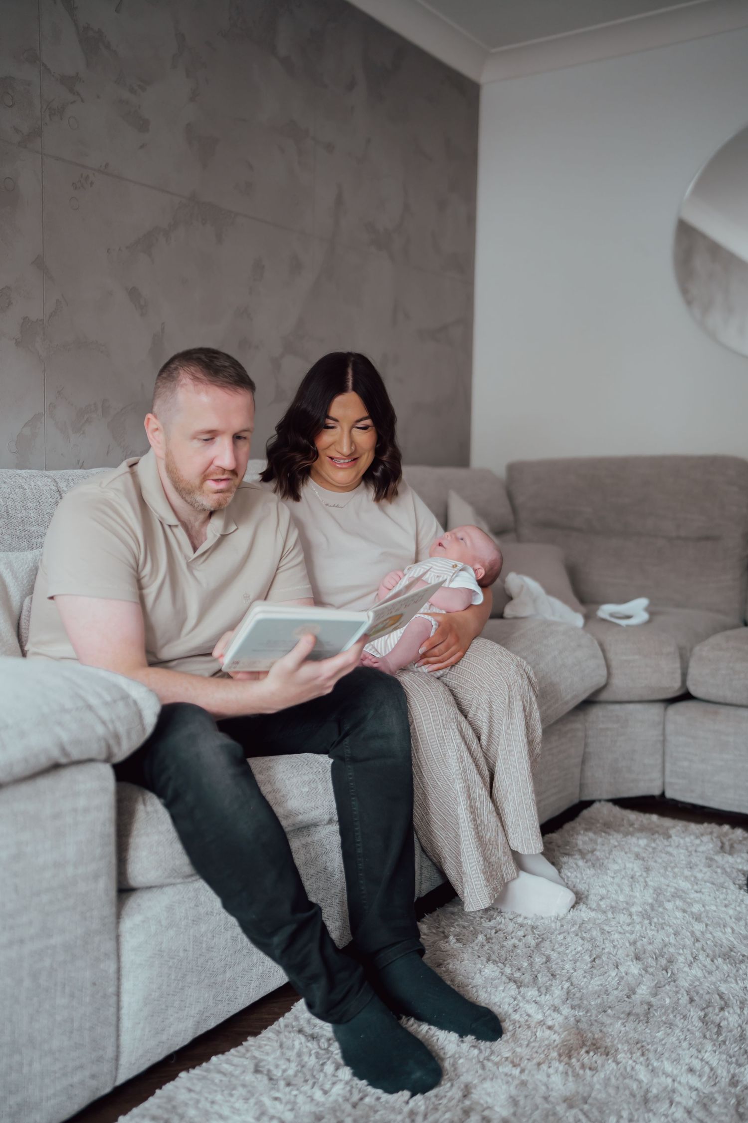 A couple sits on a gray sofa, man holding a book while woman cradles a newborn baby in a cozy living room.