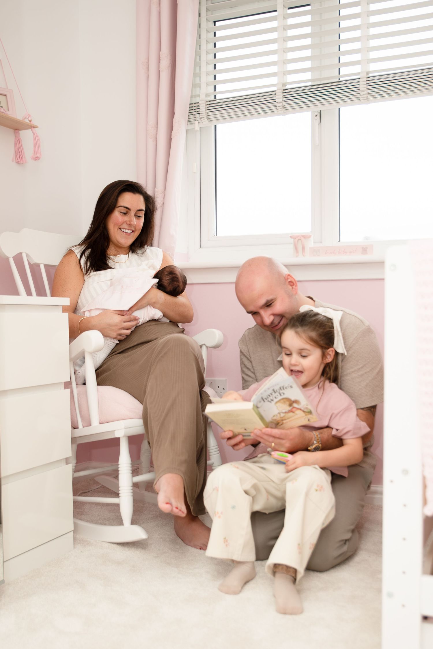 Family reading time in a cozy pink-walled room with natural light streaming through windows.