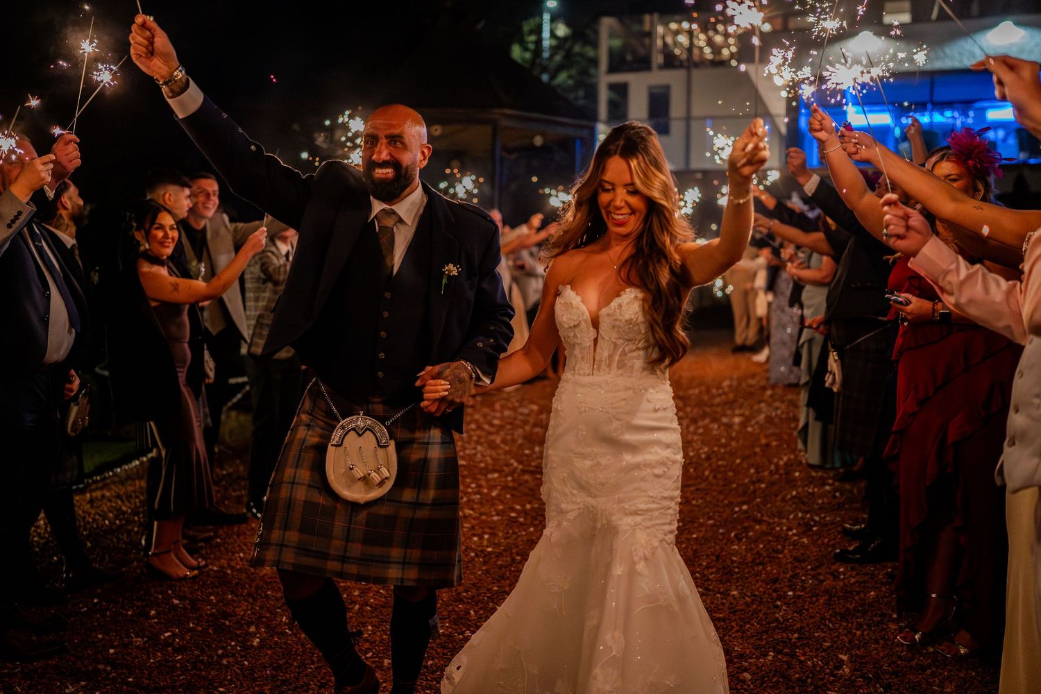 Guests toss autumn leaves at a nighttime outdoor wedding celebration as newlyweds celebrate under string lights.