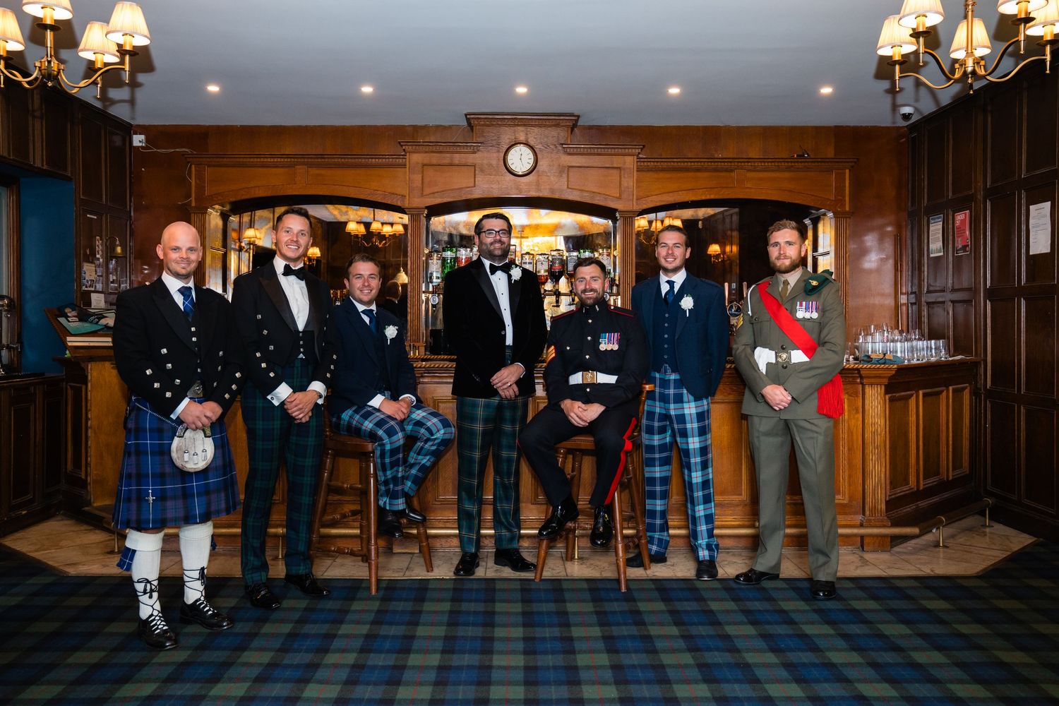 Group of men in Scottish kilts and formal attire posing together in a wood-paneled room at a wedding celebration.