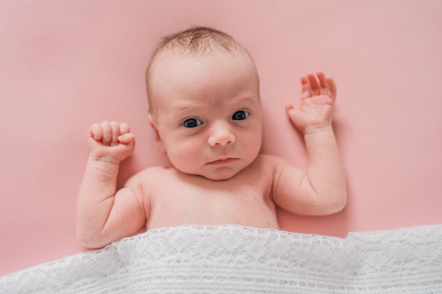 Newborn baby lying on pink background with white blanket, arms raised, looking at camera with alert expression.