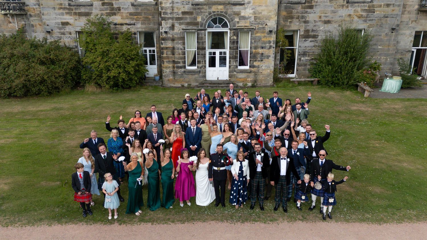 Aerial drone photo of a large wedding group gathered on green lawn outside a historic stone manor house.