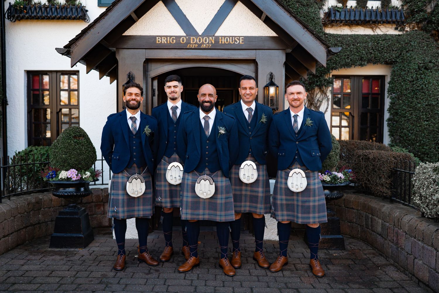 Groomsmen in matching plaid kilts and navy jackets stand together on stone steps at a Scottish wedding.