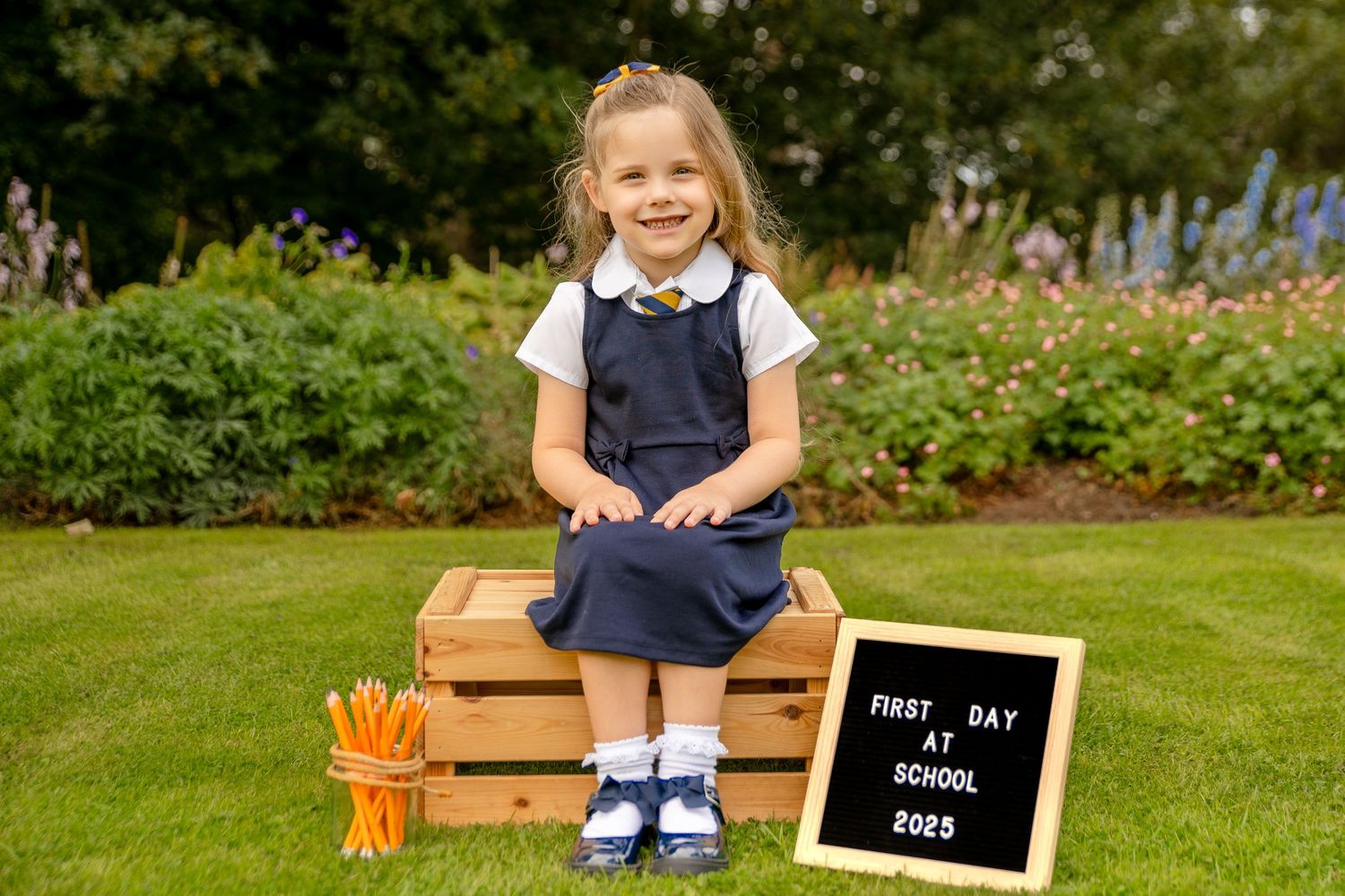 Young girl in school uniform sits on wooden crate with pencils and first day of school 2025 sign outdoors.