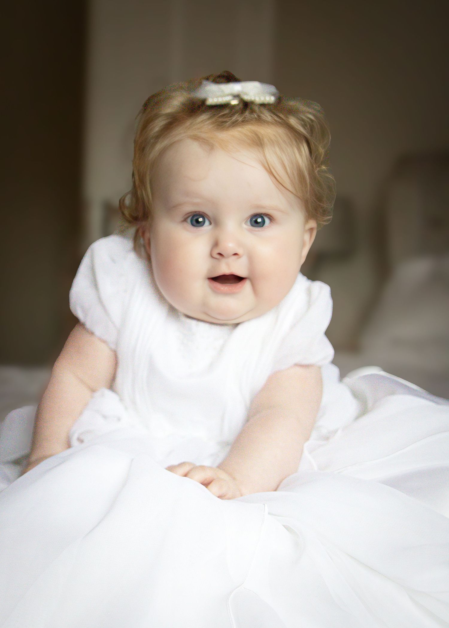 Adorable baby girl with blue eyes wearing a white dress and bow headband, smiling sweetly at the camera.