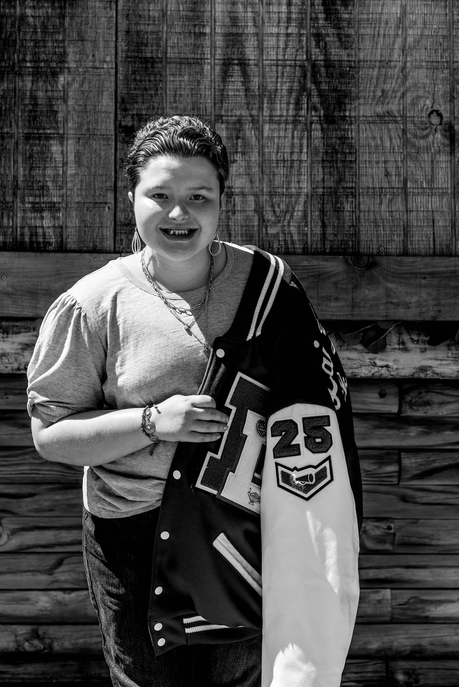 A person poses with a varsity letterman jacket against a rustic wooden wall in black and white.